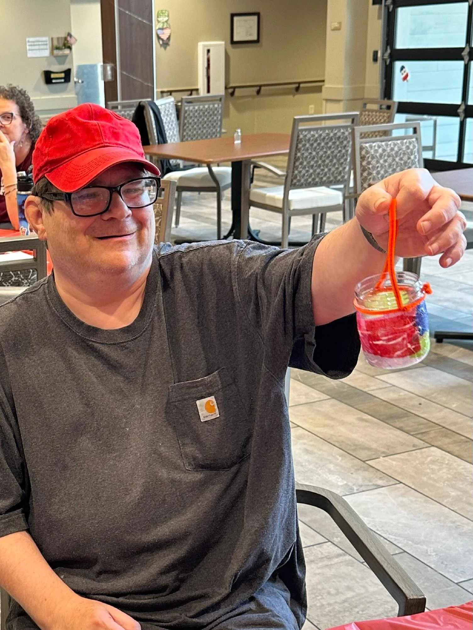 A man wearing glasses and a red cap smiling and holding a colorful jar with a bright orange straw inside, sitting at a table in a casual indoor setting.