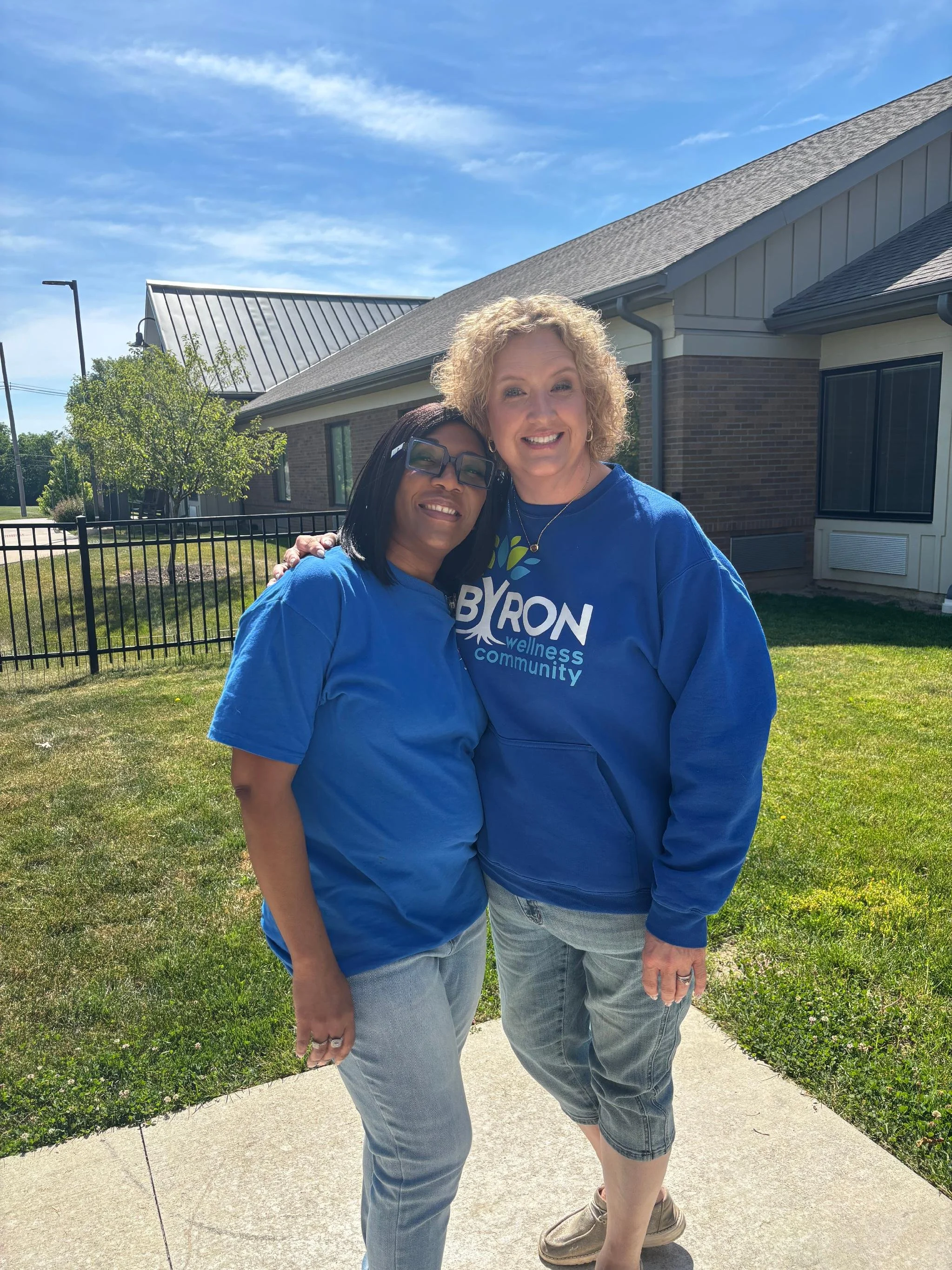 Two women embracing outdoors on a sunny day, in front of a residential building with a manicured lawn, one wearing glasses and both wearing blue shirts.