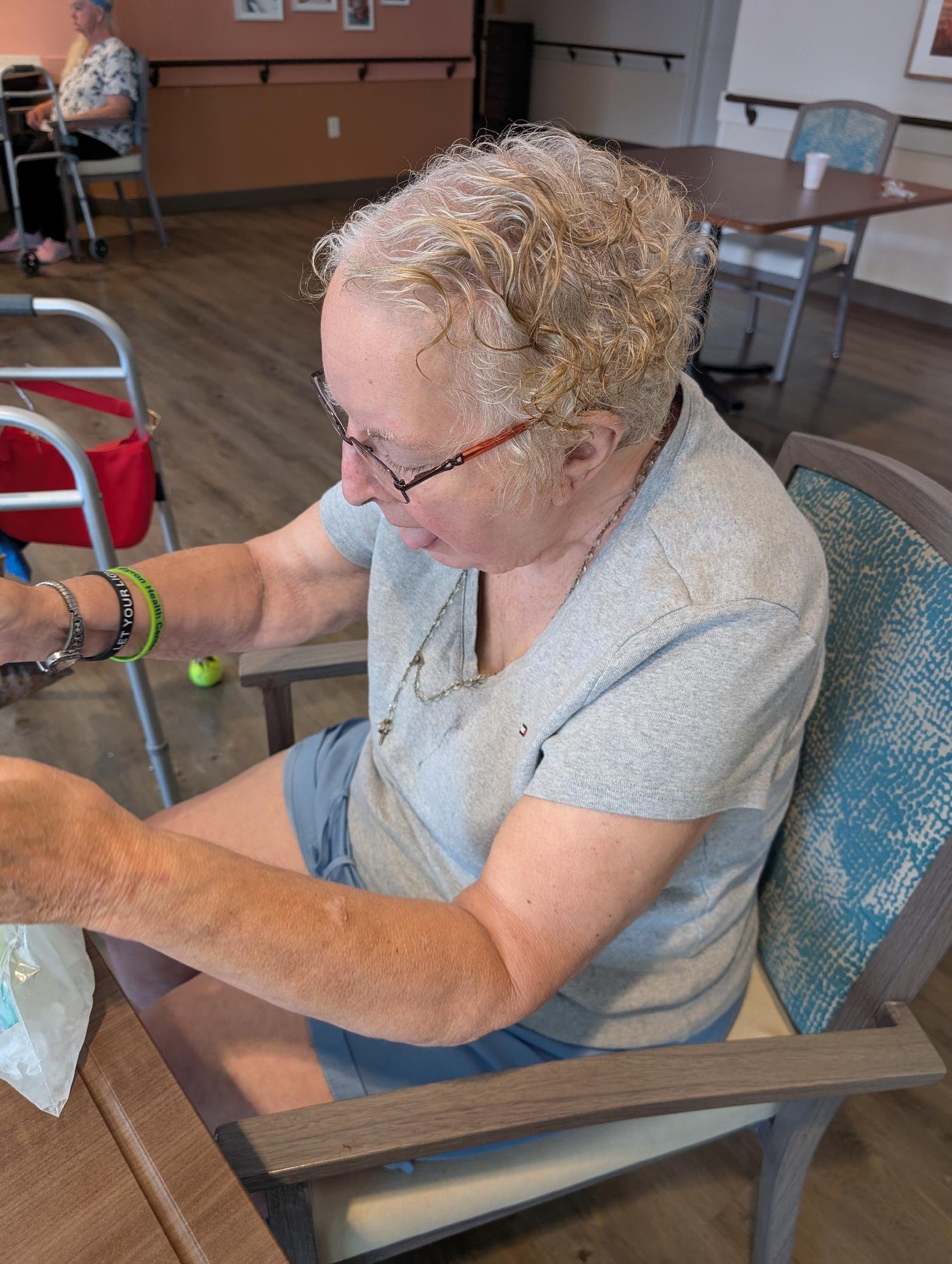 An elderly woman with curly blonde hair, glasses, wearing a gray t-shirt and blue shorts, sitting at a wooden table in a communal room. She appears to be engaging with items on the table. In the background, another person with a walker is seated near