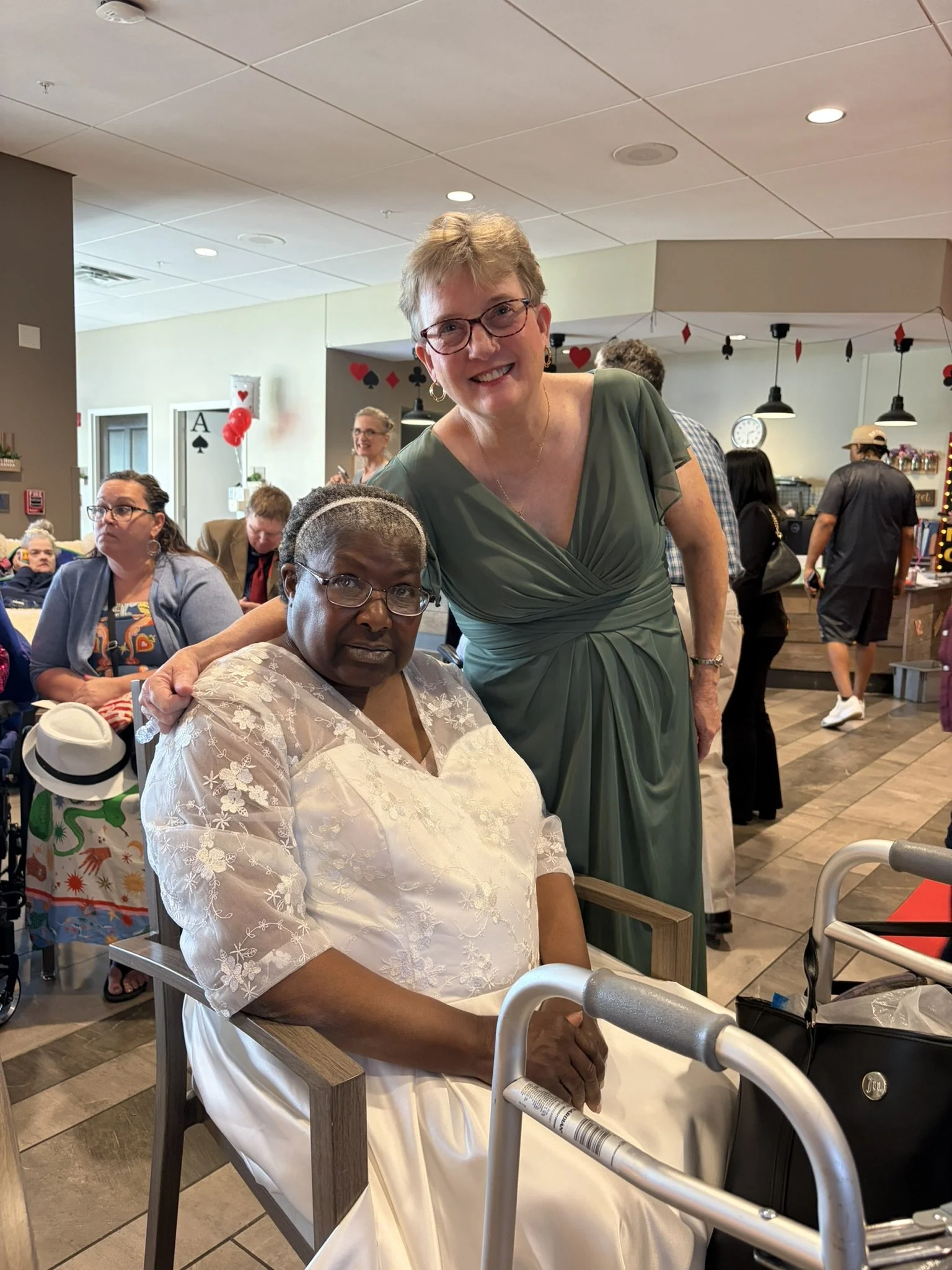 A woman in a white dress sitting in a restaurant with a walker in front of her, another woman in a green dress standing next to her, and people in the background dining and walking around.