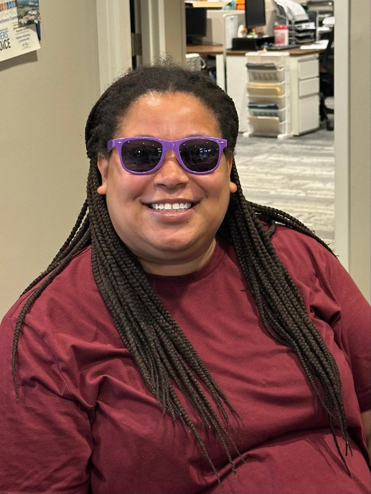 A smiling woman with long braided hair wearing purple sunglasses and a maroon shirt, sitting inside an office or workspace.