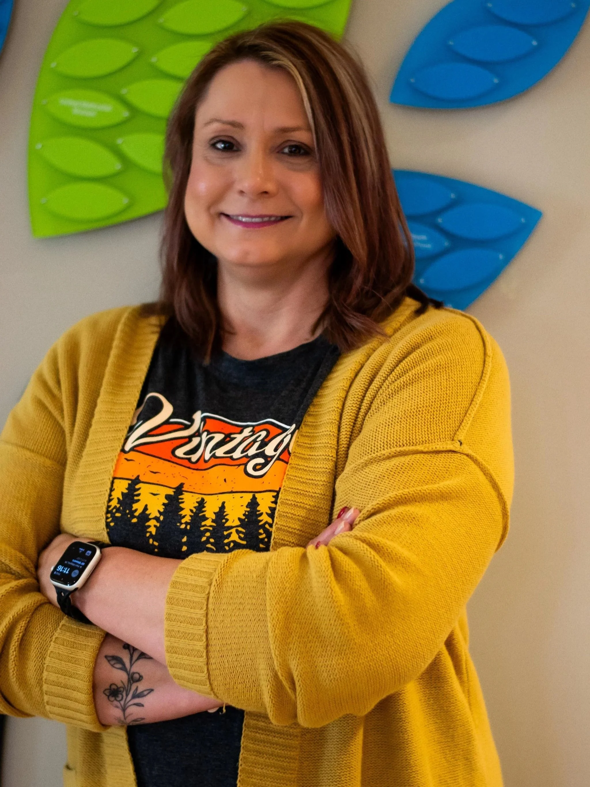 A woman with shoulder-length brown hair wearing a yellow cardigan, black T-shirt, and an Apple Watch, standing in front of a wall decorated with colorful, flower-shaped art pieces in green and blue.