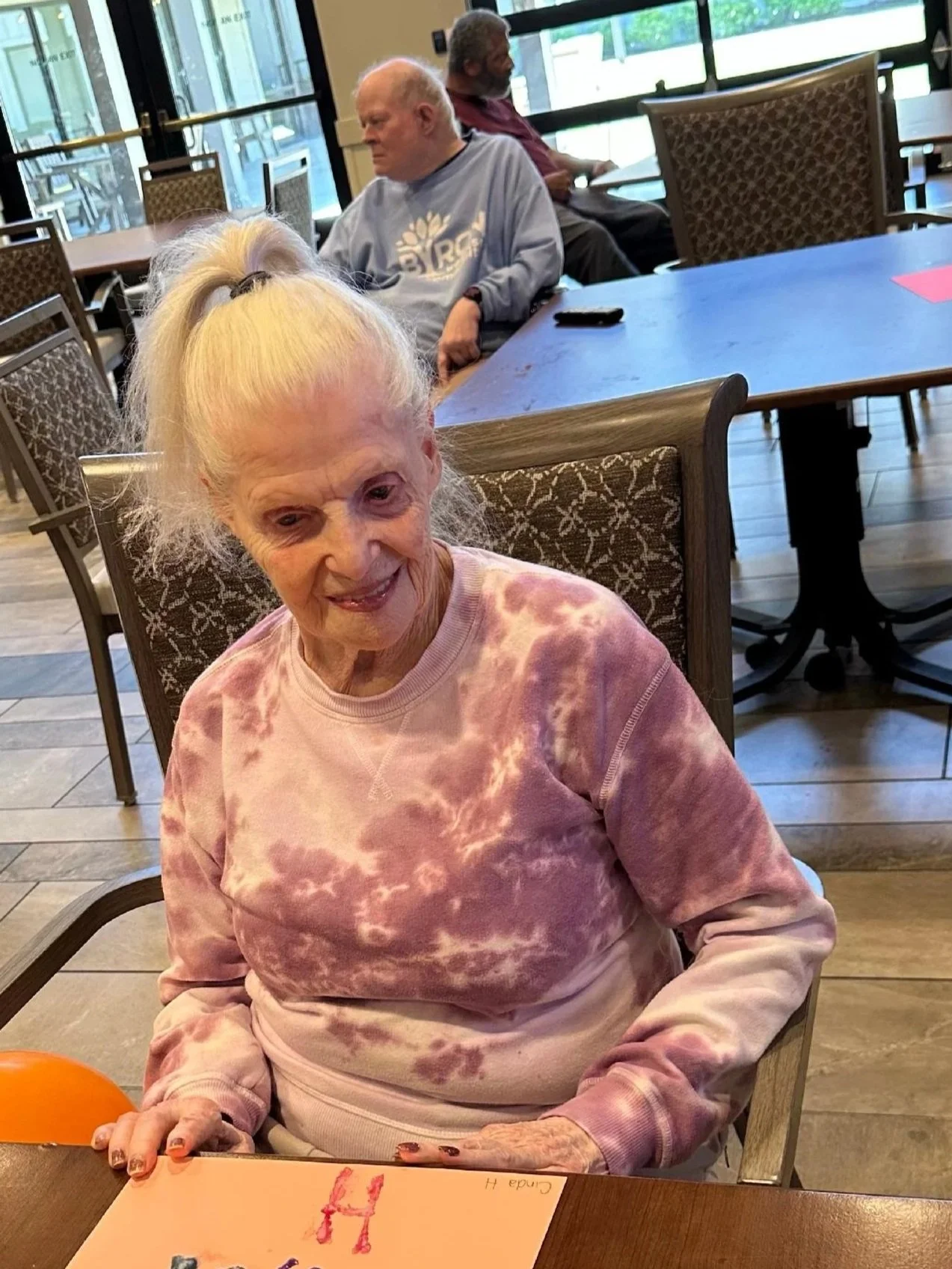 An elderly woman with long blonde hair in a ponytail, wearing a pink tie-dye sweatshirt, smiling and sitting at a table in a restaurant or cafe.