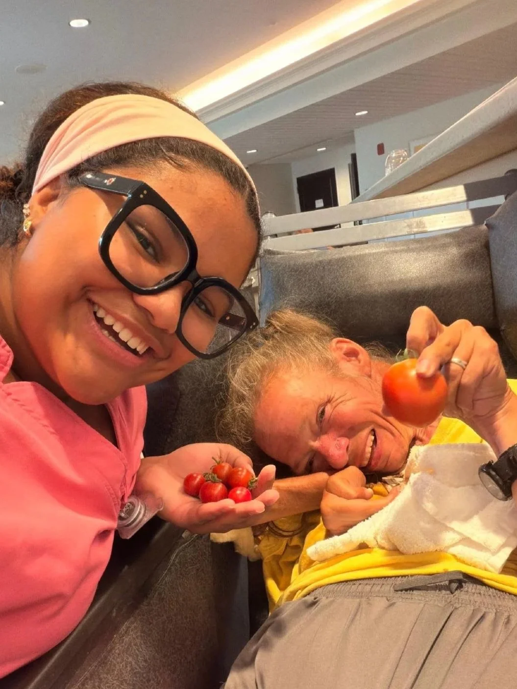 Young woman smiling and posing with an elderly person lying on a couch, holding a tomato, in an indoor setting.