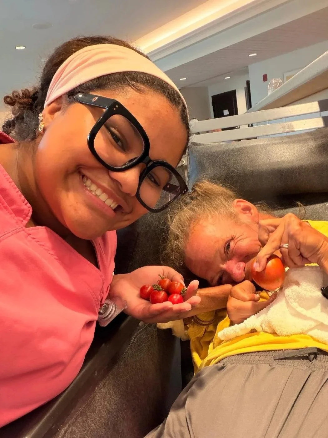 A young woman with glasses and a pink shirt smiling at the camera while holding cherry tomatoes. An elderly woman with curly hair lies on a bed, holding a tomato and biting it, smiling and holding cherry tomatoes.