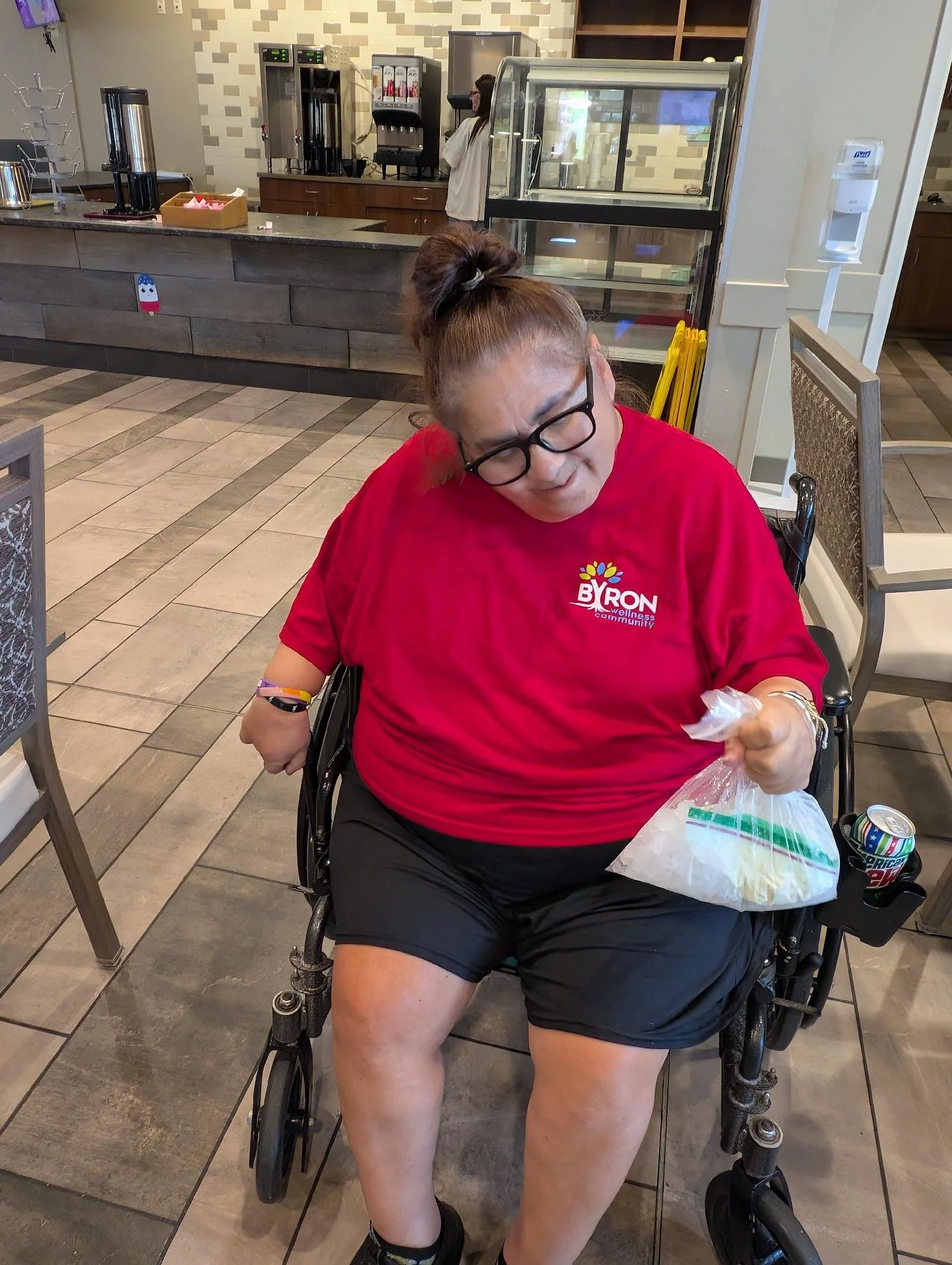 A woman in a wheelchair wearing glasses and a red shirt with a logo, sitting in a casual dining area, holding a plastic bag with what appears to be food. The background shows a counter with a coffee machine, a woman in white working behind the counte