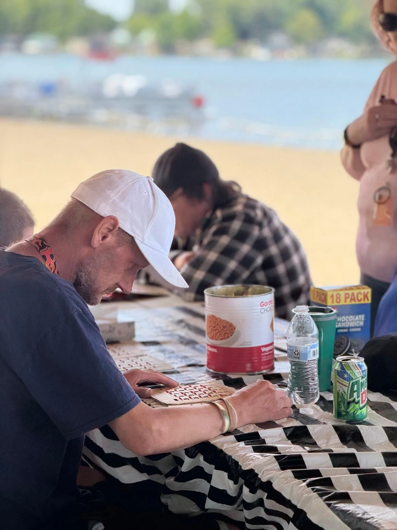 People sitting at a table outdoors, some with food and drinks, near a body of water with boats. The table has a black and white checkered tablecloth.