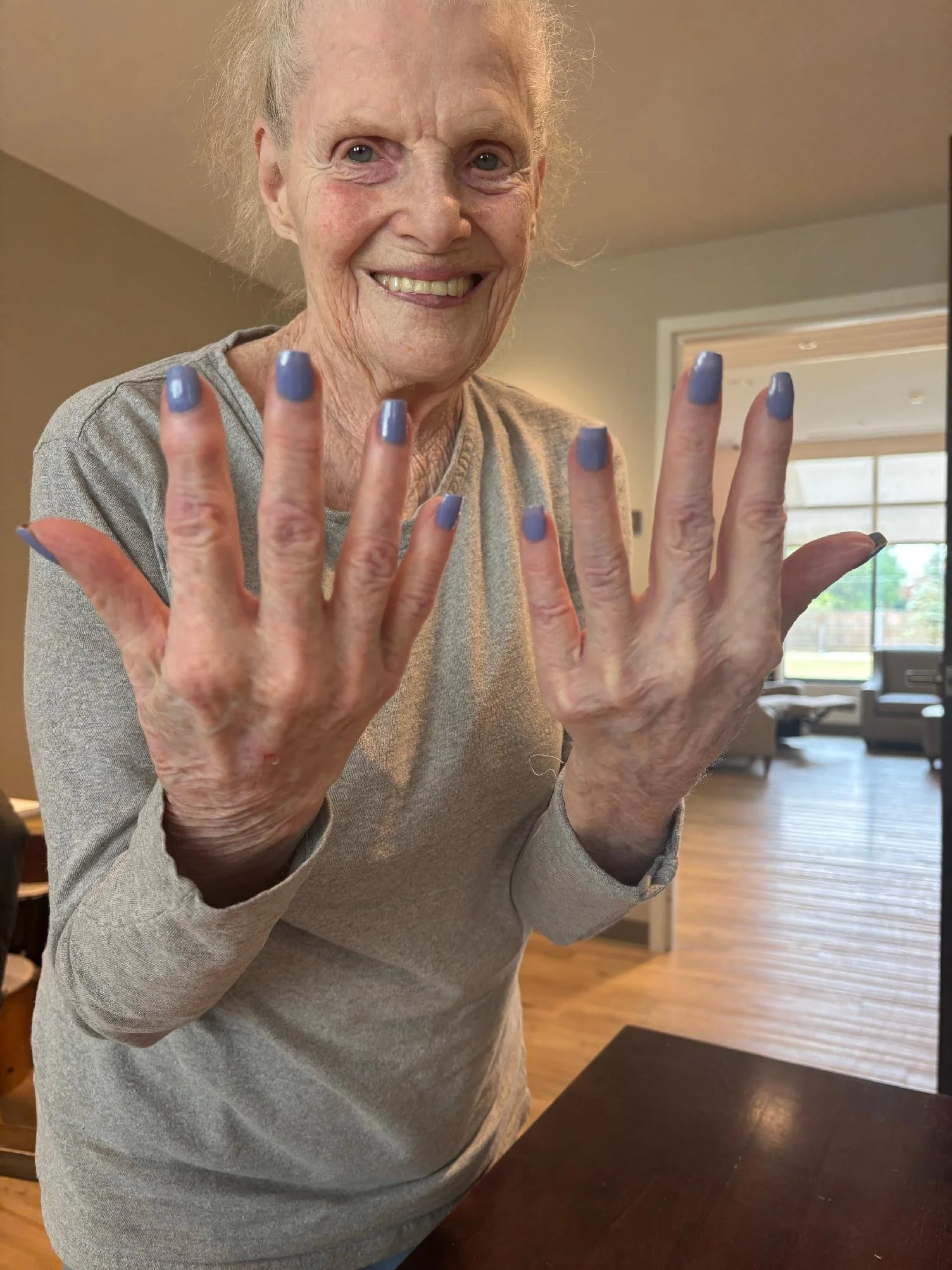 An elderly woman with gray hair smiling and showing off her freshly painted purple nails inside a cozy home with wooden floors and large windows.