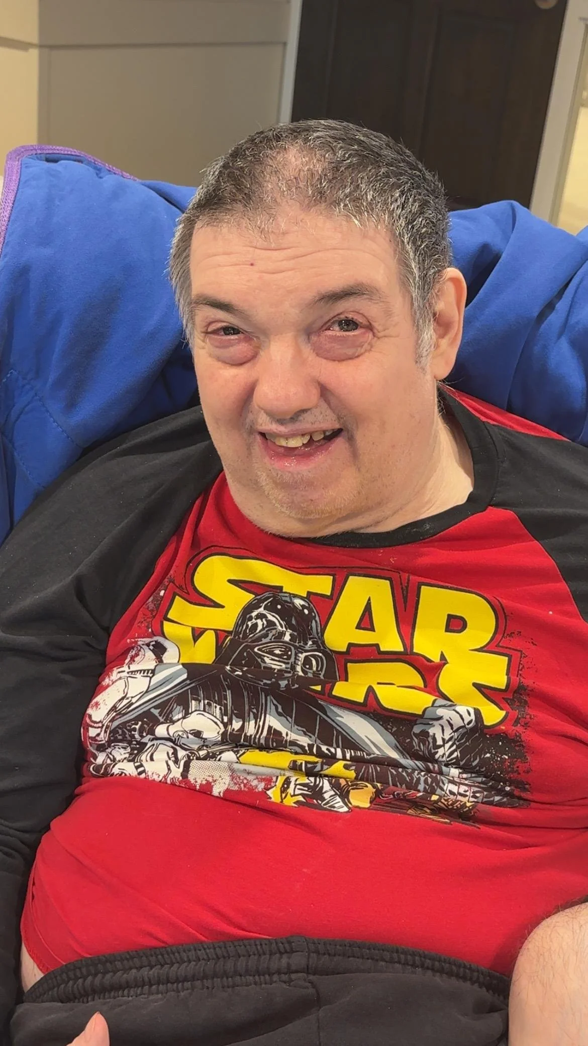 A smiling man with short gray hair, wearing a red Star Wars t-shirt, sitting on a black couch with a blue blanket behind him in a room with beige walls.