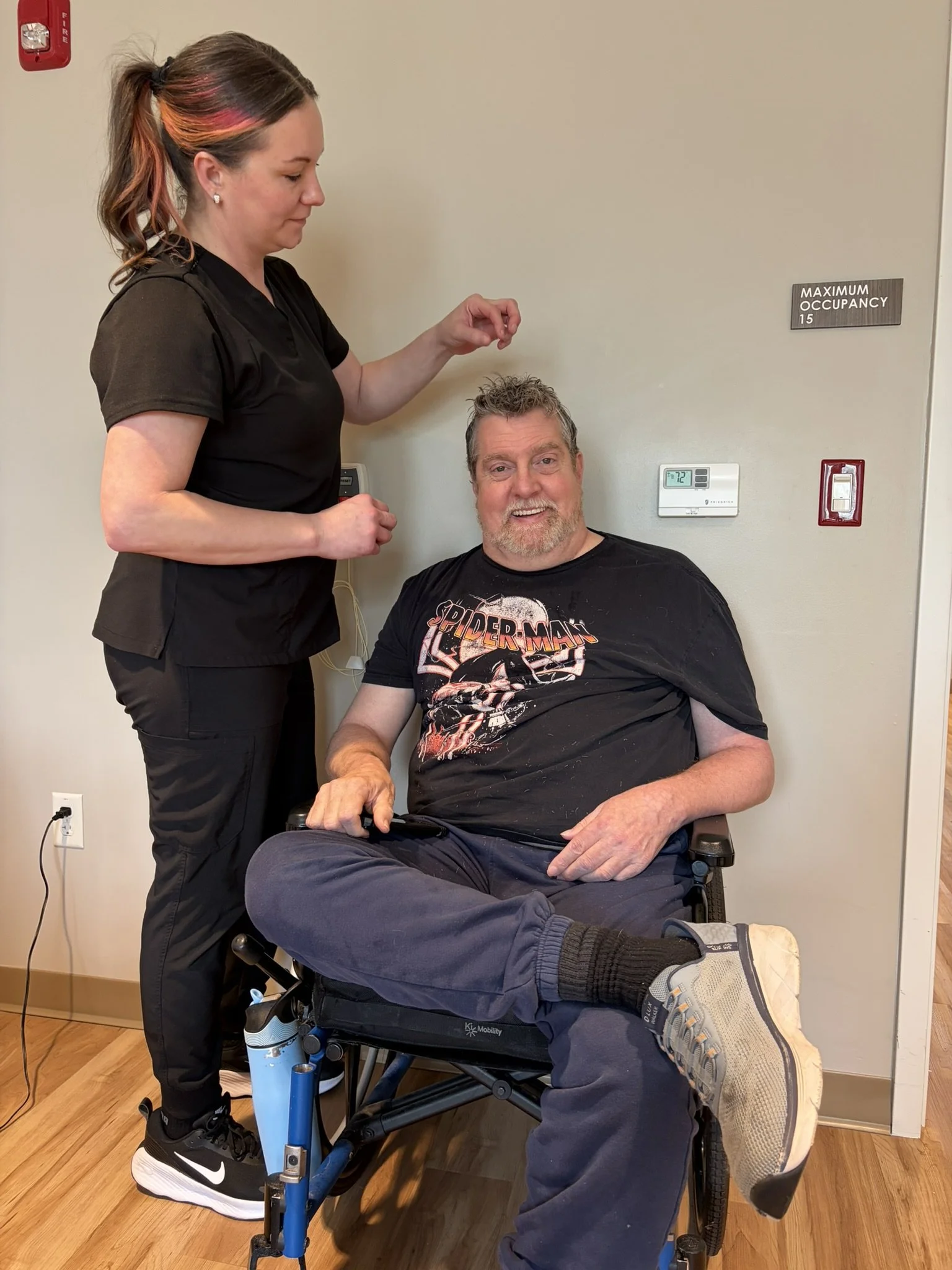 A man in a wheelchair smiling, wearing a Spider-Man T-shirt and gray pants, is being attended to by a woman standing next to him, in a room with a wall-mounted thermostat and a fire alarm. The woman has long hair with pink streaks and is dressed in b