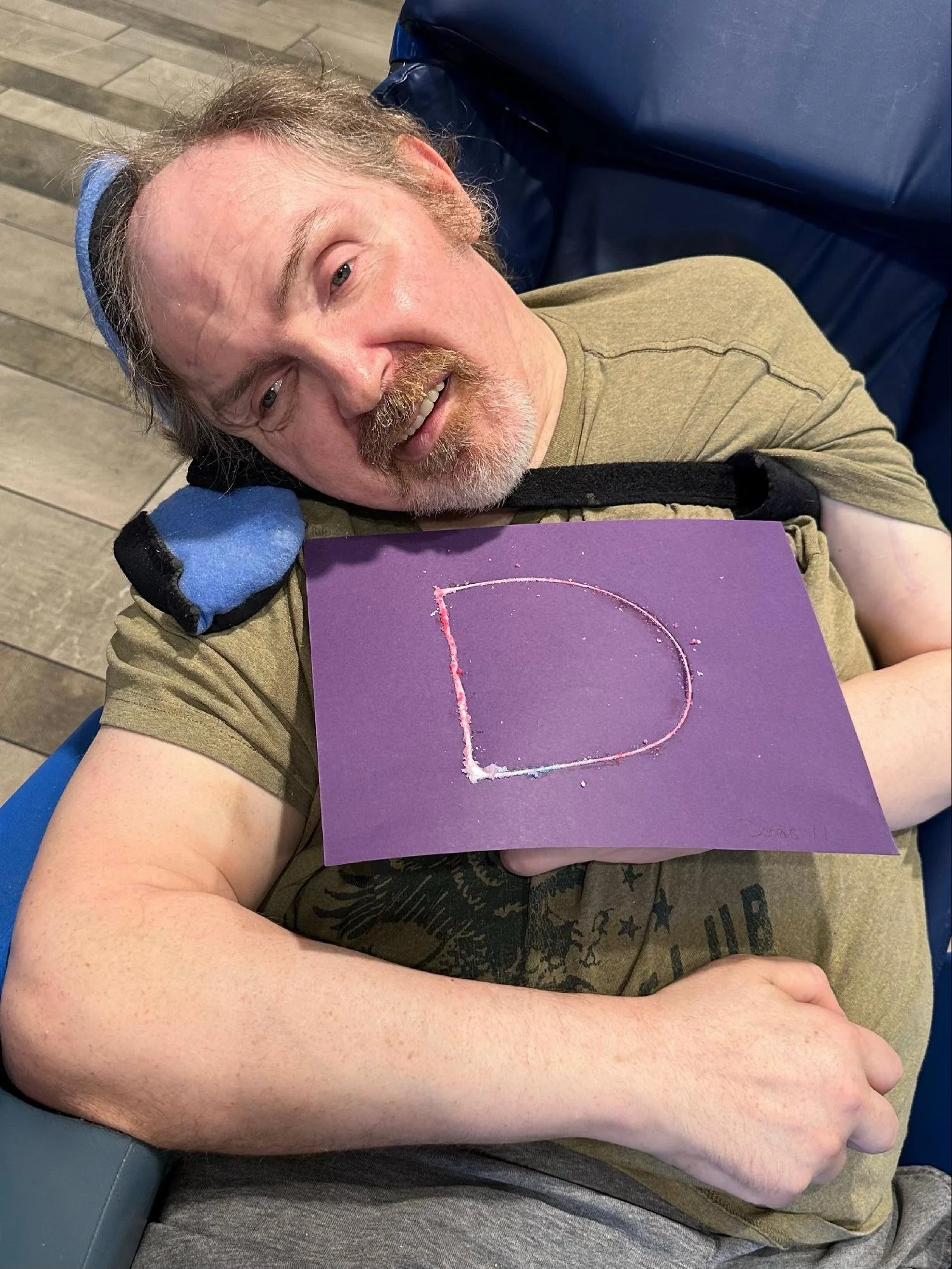 A man with a beard and mustache lying down on a chair with his head resting on a blue neck pillow. He has a relaxed expression and is holding a purple card with the letter 'D' drawn in pink. The man is wearing a green T-shirt and has light complexion