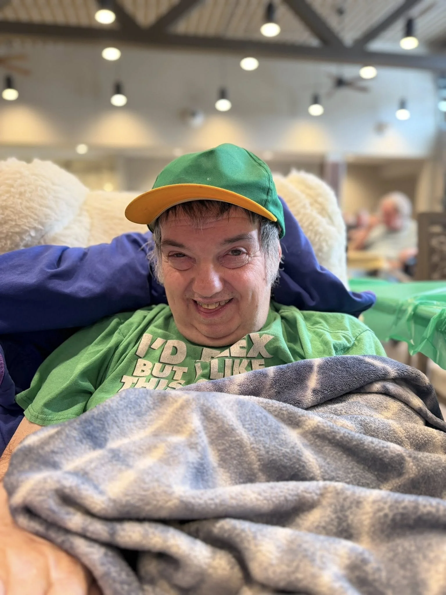 A smiling man lying on a table in a restaurant with a green cap and a gray shirt. The background shows blurred patrons and restaurant interior lights.