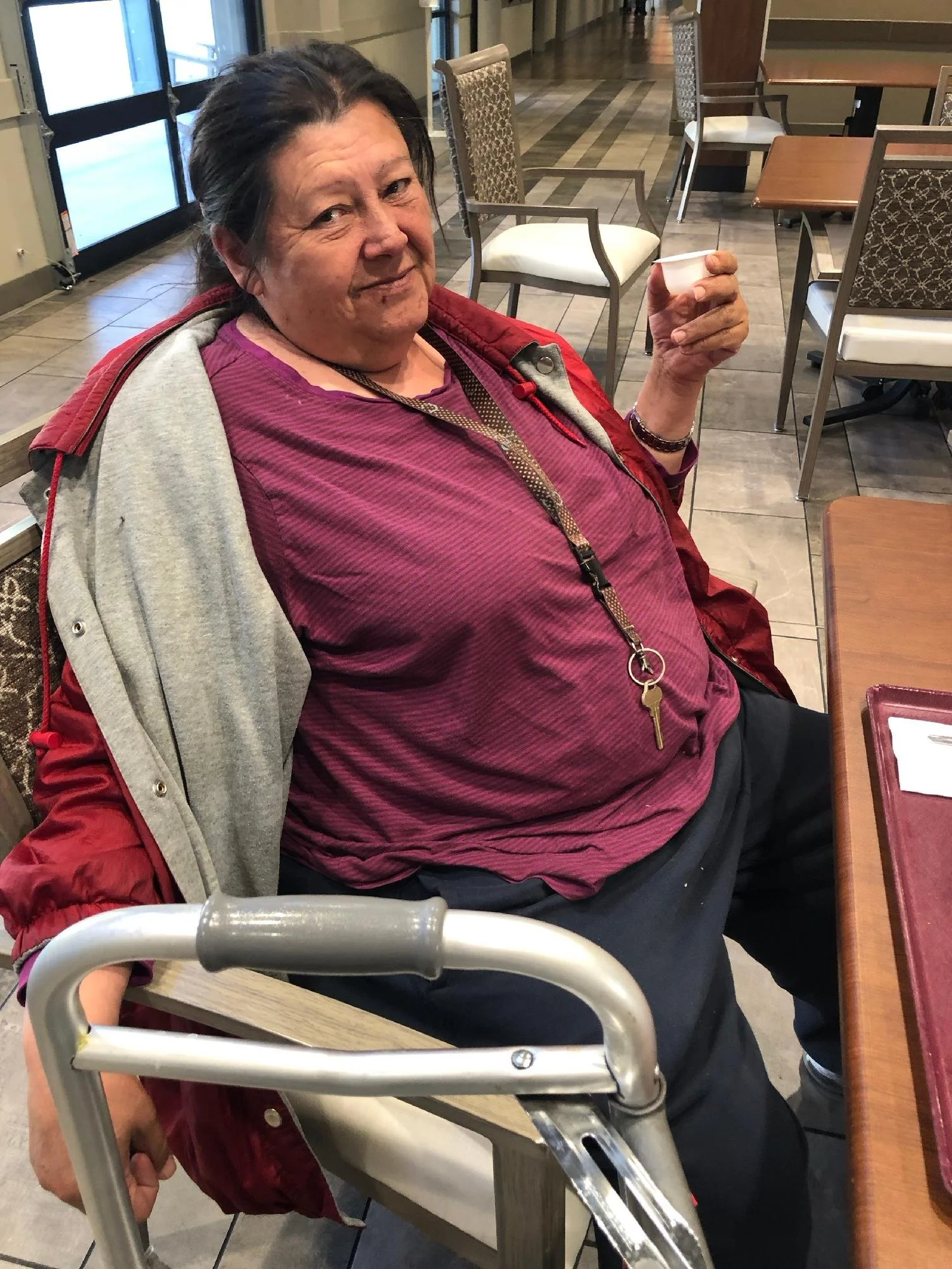 An elderly woman sitting at a restaurant table, holding a small white cup, with a walker in front of her and empty chairs and tables in the background.
