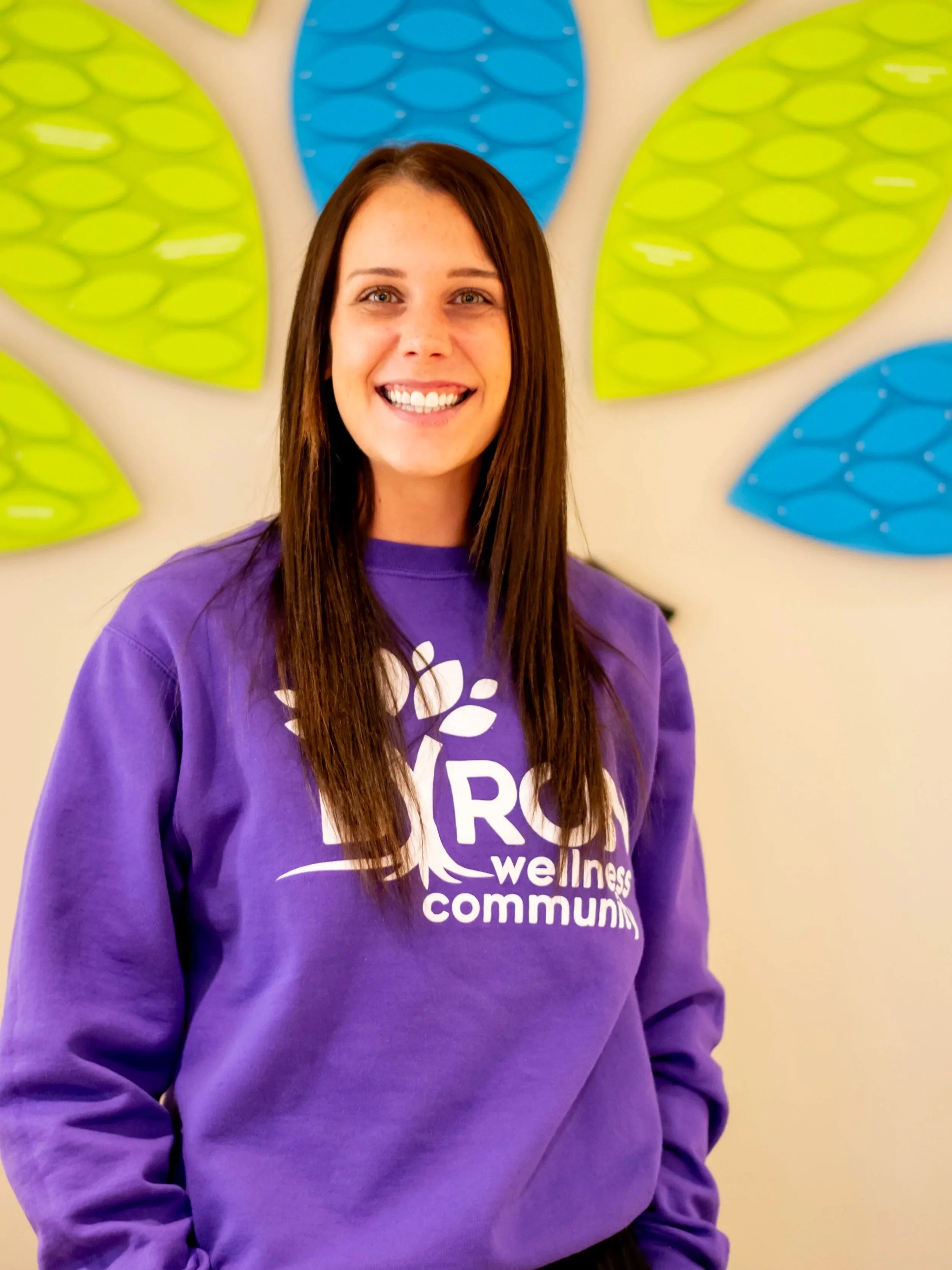 A woman with long brown hair smiling, wearing a purple sweatshirt with white text that reads 'Wellness community'. She is standing in front of a colorful wall art featuring large green and blue leaves.