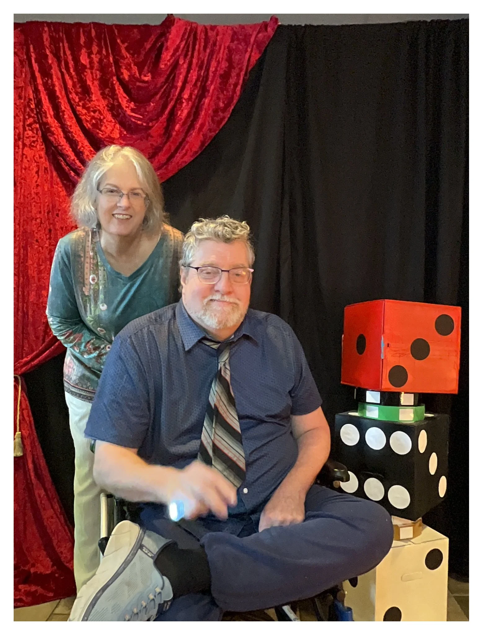 Two smiling people, one woman with silver hair and glasses standing behind a man sitting in a wheelchair, in front of red and black curtains. There is a large red and black dice-shaped lamp on a small table next to them.