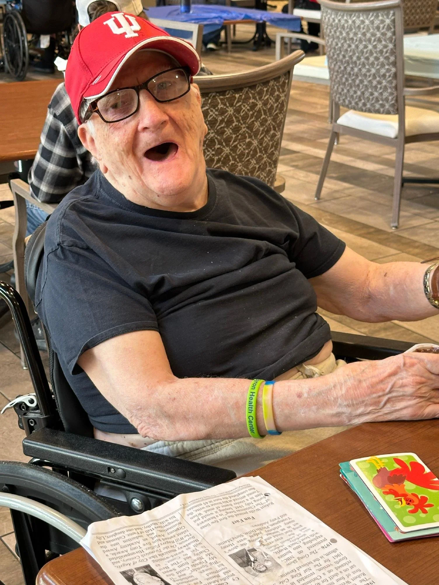 An elderly man in a wheelchair wearing a red Indiana University baseball cap, glasses, and a black t-shirt, sitting at a dining table with a newspaper and colorful children's books, in a dining area with wooden floors and patterned chairs.