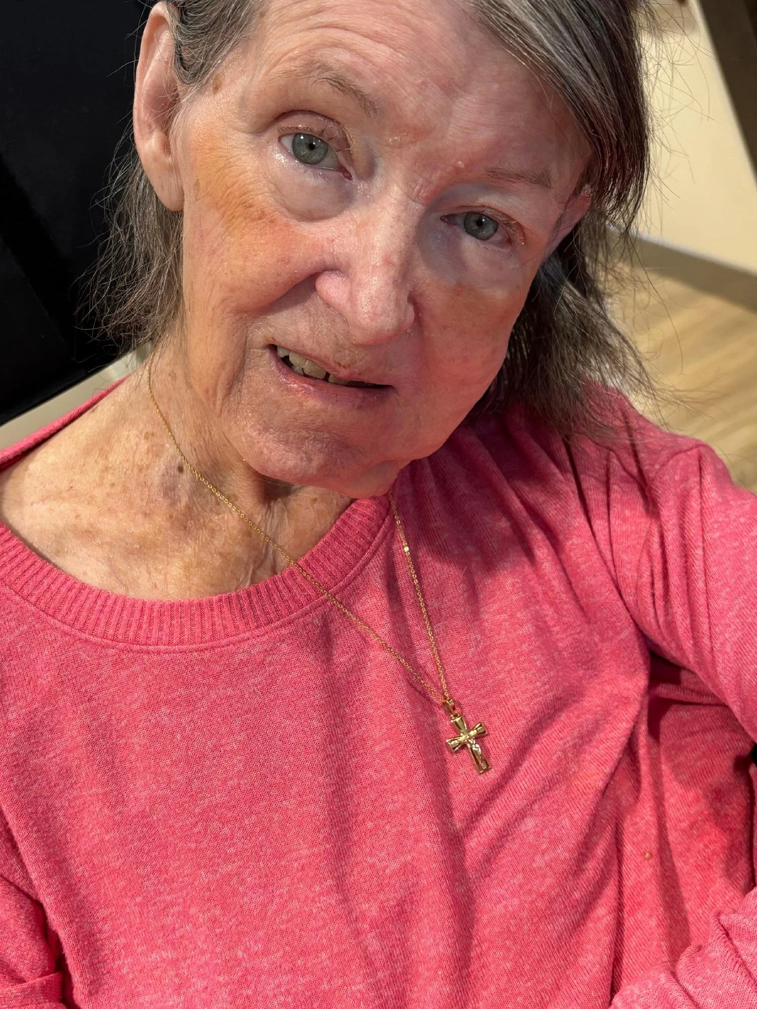 Close-up of an elderly woman with gray hair and blue eyes, wearing a pink shirt and a gold necklace with a cross pendant, smiling softly.