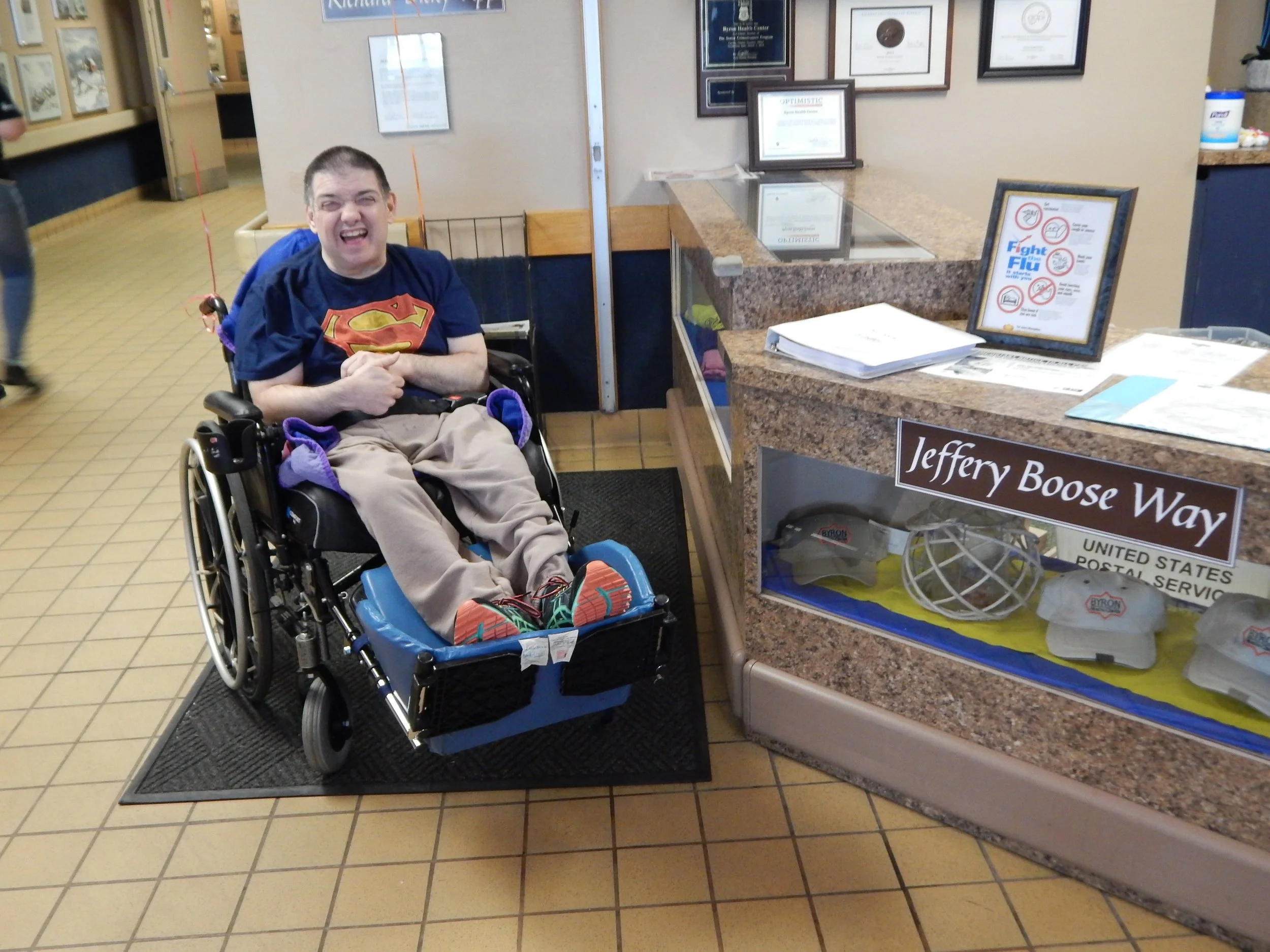 A young man in a wheelchair wearing a Superman T-shirt, smiling in a reception area with framed awards and a sign about fighting flu on the counter.