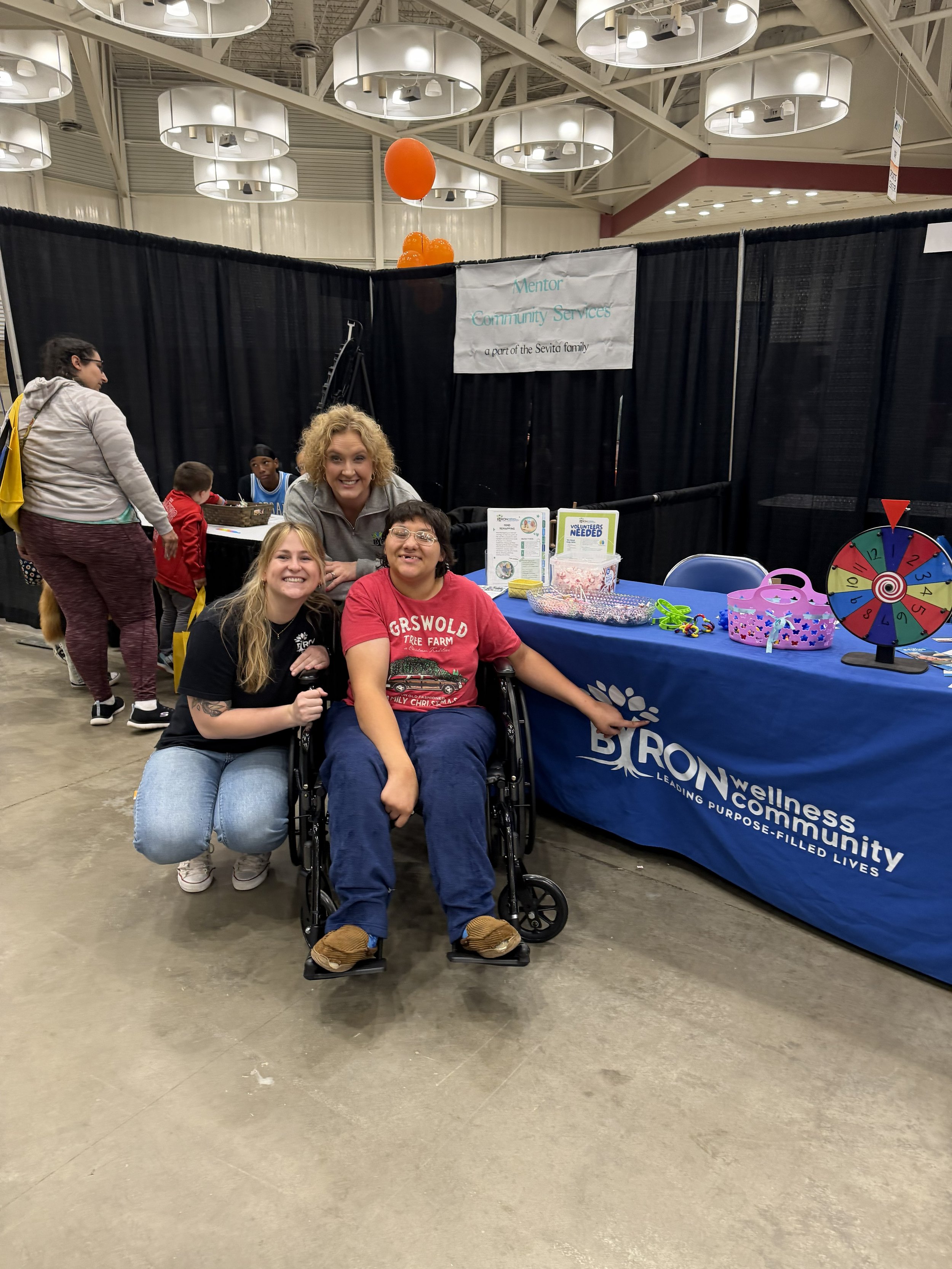 Smile of a woman and a person in a wheelchair with two women behind them at a community event booth with a sign that reads 'Mentor Community Services, a part of the Sevita family.' Other people are standing and sitting nearby.