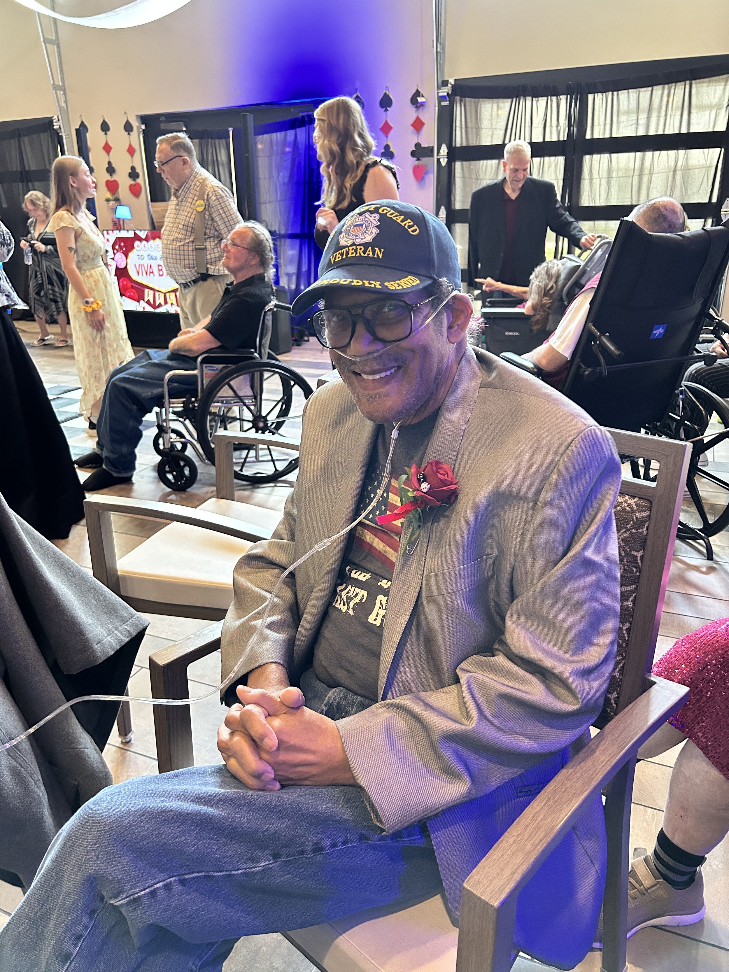 A group of elderly people and a young woman celebrating at an indoor party. Some are in wheelchairs, others seated or standing, with decorations resembling playing card suits. One man wears a cap labeled "VETERAN" and is smiling. The atmosphere is fe
