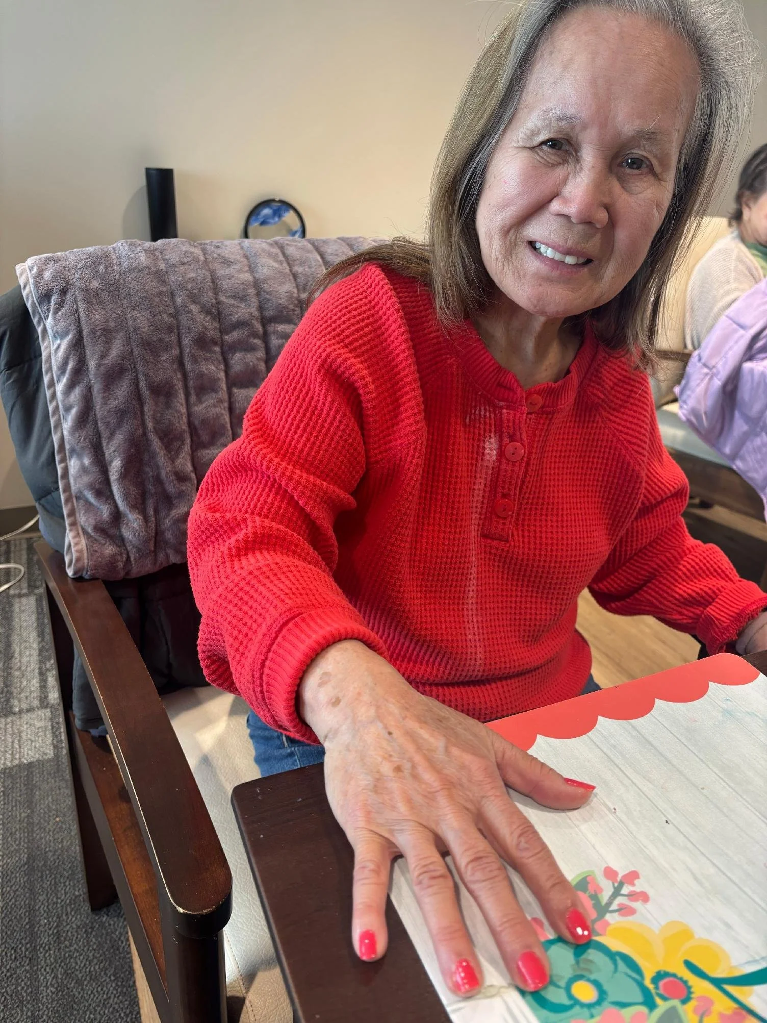 An elderly woman with gray hair and a red sweater sitting at a table, stretching her hand across a colorful greeting card with floral designs. She is smiling and wearing bright red nail polish.