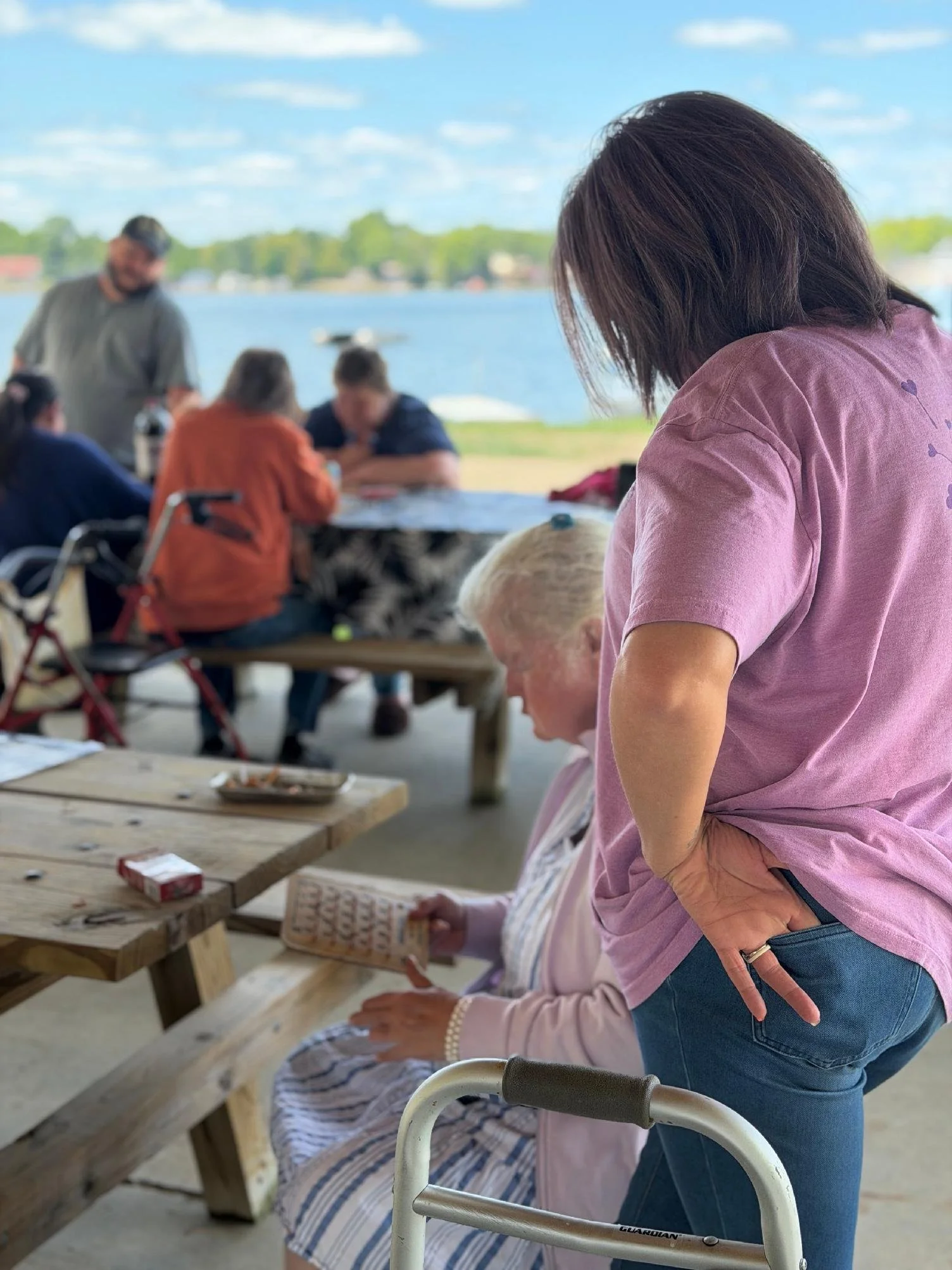 People gathered outdoors by a lake, with a woman in a pink shirt and blue jeans standing next to an elderly woman seated at a wooden picnic table. The scene features a scenic lake view with trees and houses in the background.