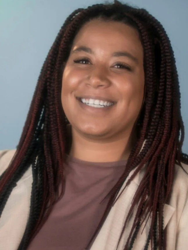 A woman with long braids smiling at the camera, wearing a light-colored blazer and a mauve shirt, against a blue background.
