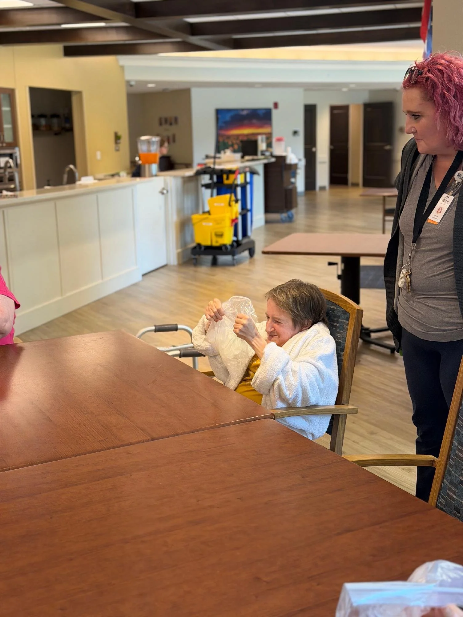 An elderly woman sitting in a chair at a dining table, holding a plastic bag and being assisted by a woman standing nearby, in a dining area of a care facility or restaurant.