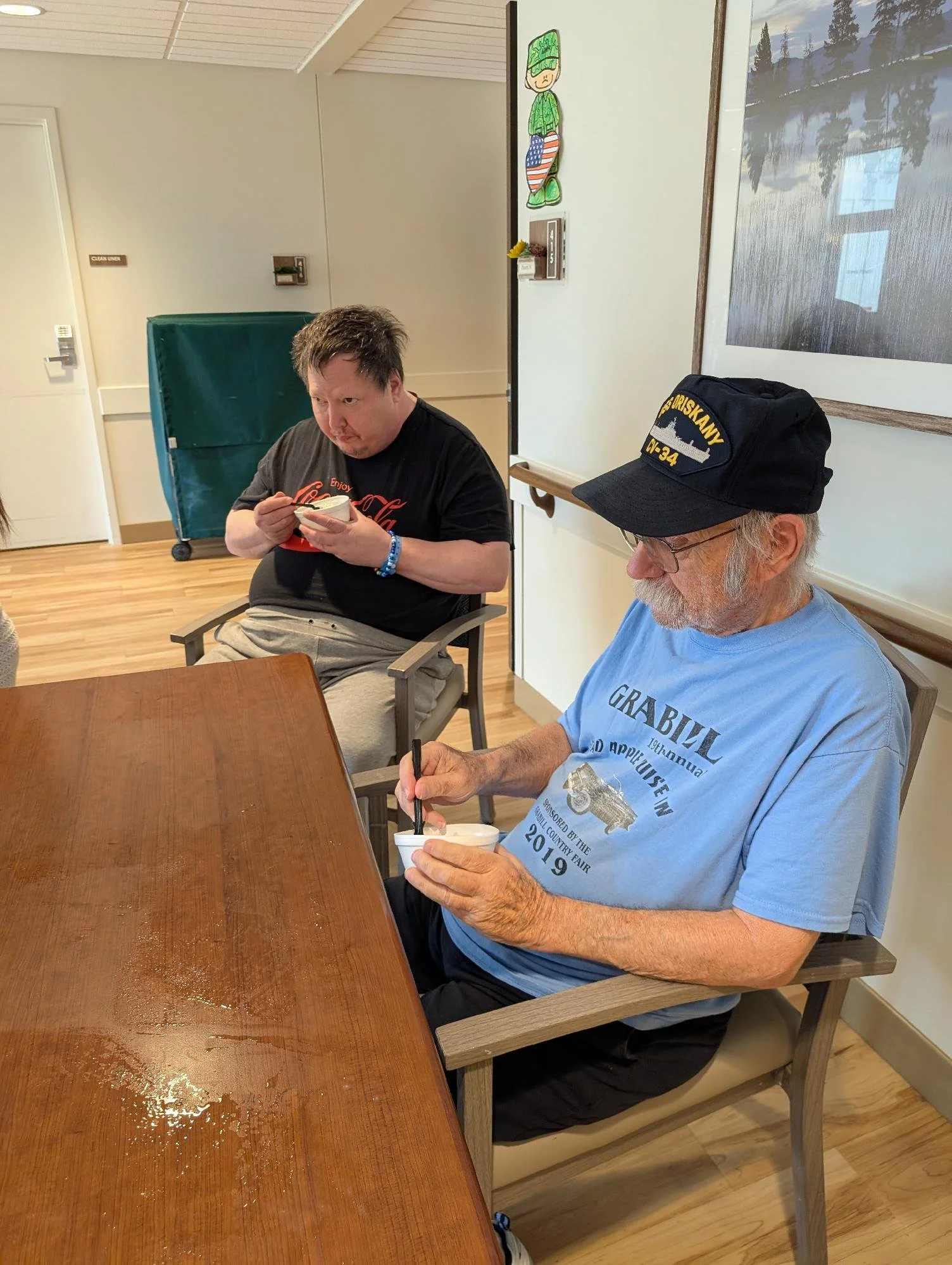 Two men, one with gray hair and glasses wearing a navy blue cap, and the other with messy dark hair, sitting at a wooden table and eating ice cream in a room with wooden flooring and light-colored walls. The man with the cap is wearing a light blue s