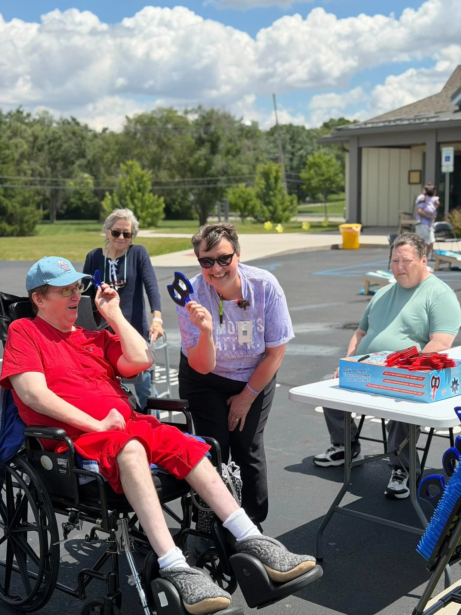 A group of people gathered outdoors in a parking lot on a sunny day, with some sitting and others standing. There are trees, a building, and blue sky with clouds in the background. One person in a wheelchair, wearing a red shirt, is being handed a pa