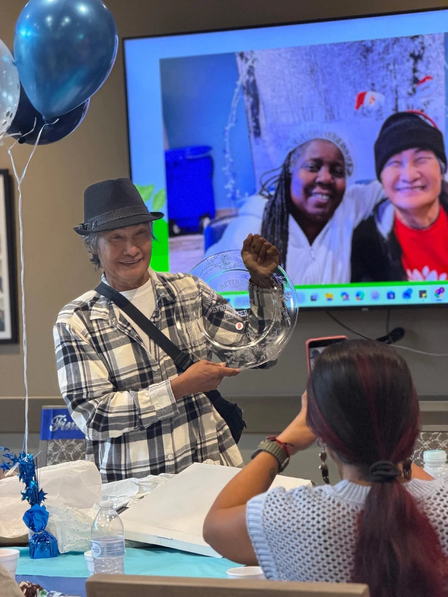 An elderly woman wearing a plaid shirt and hat is holding a glass sculpture and smiling at a celebration. A woman with long hair and another with short hair are sitting at the table. In the background, a large TV screen displays a photo of two women 