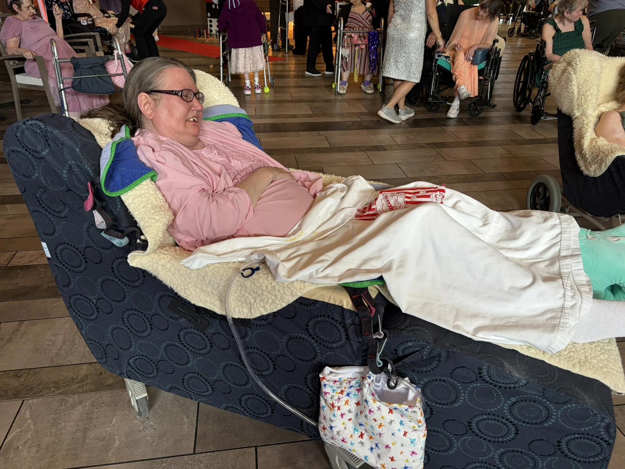 A woman in a wheelchair reclines with her eyes closed and smiling, in a room with other women in wheelchairs and children, some standing or sitting. The woman is wearing glasses and a pink top, with a blanket over her lap, and appears relaxed.