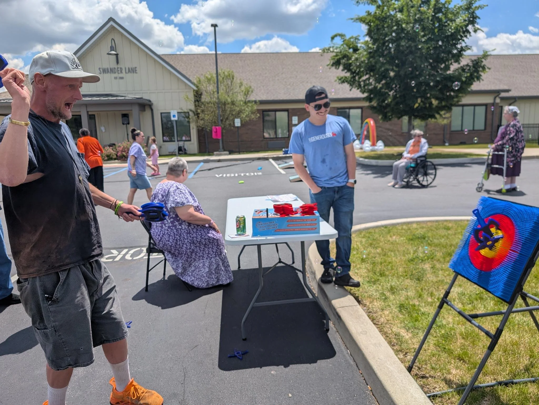 A group of people outdoors in a parking lot with a building labeled 'SWANDER LANE' in the background. Two men are wearing casual clothes and appear to be having fun, one raising his fist and shouting, the other smiling and wearing sunglasses. There a