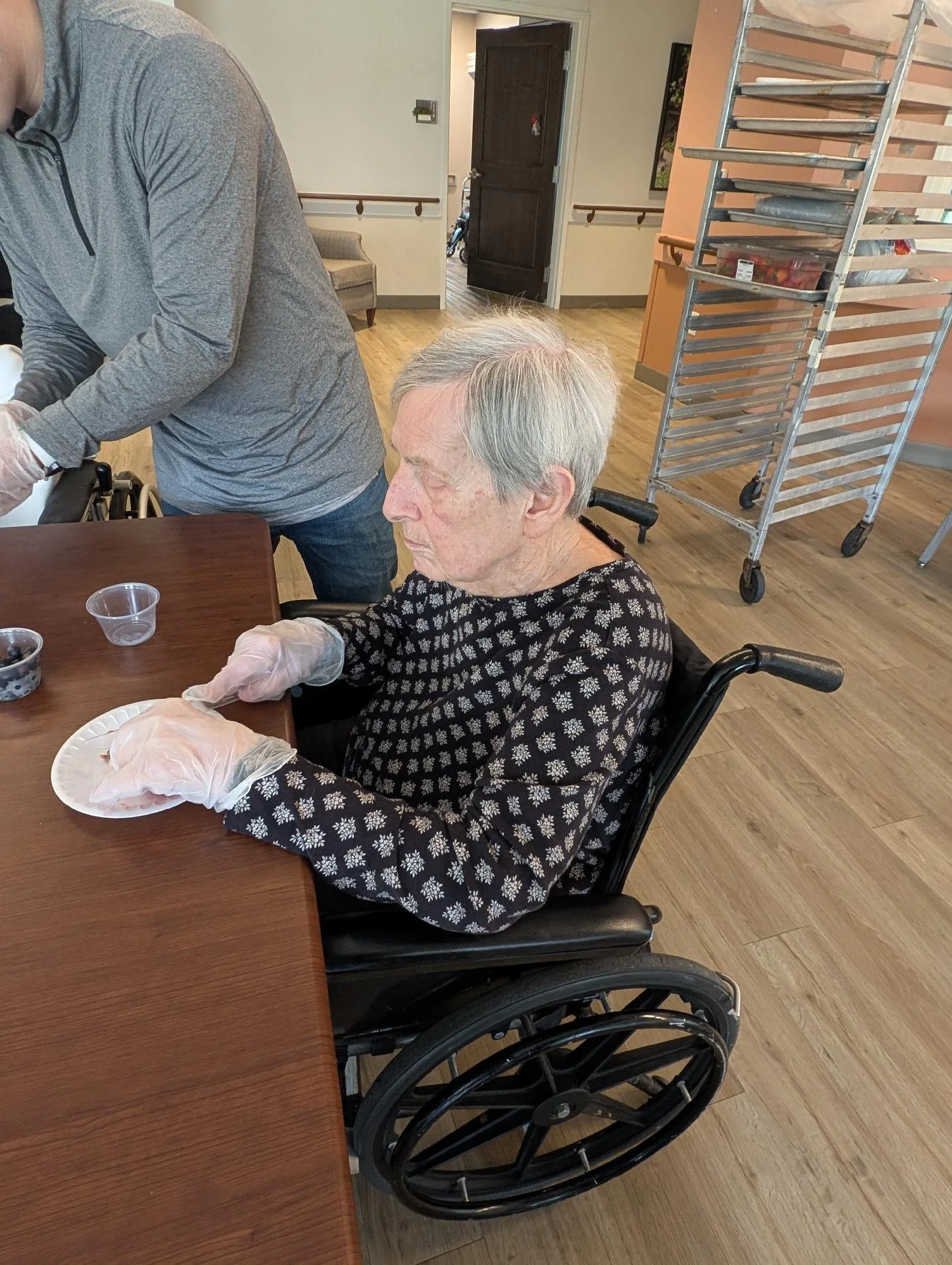 An elderly woman with gray hair in a wheelchair, wearing a black and white floral blouse, sits at a table with a paper plate and small cups, while a caregiver in gray long-sleeve shirt assists her in a room with wooden flooring, a metal cart, and a d