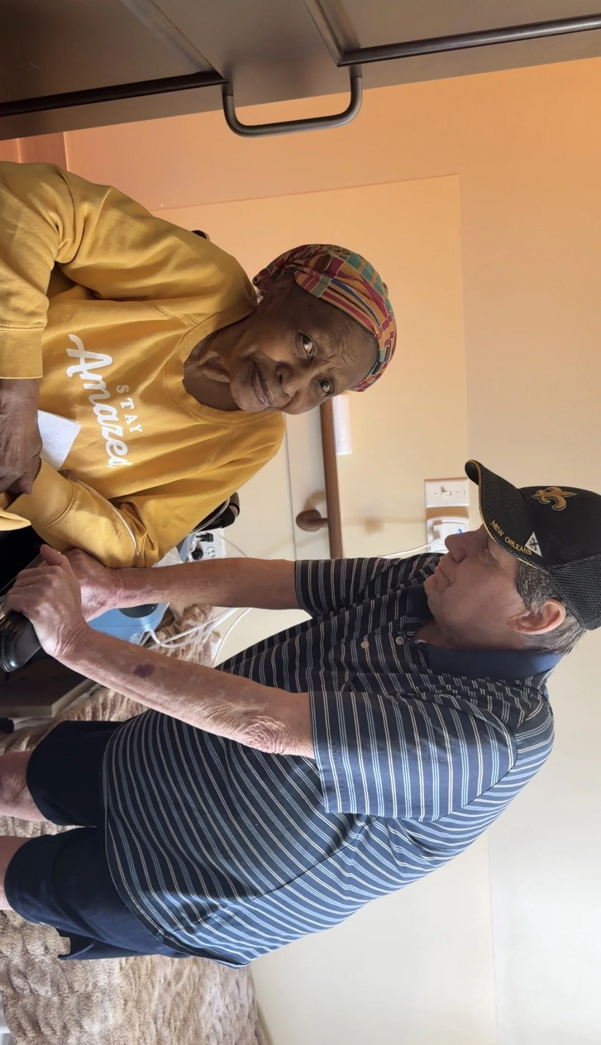 An elderly woman with gray hair wearing a colorful headscarf, a yellow sweatshirt, and tear-stained face sitting in a wheelchair in a room, chatting with a man in a baseball cap, striped polo shirt, and shorts who is leaning towards her.