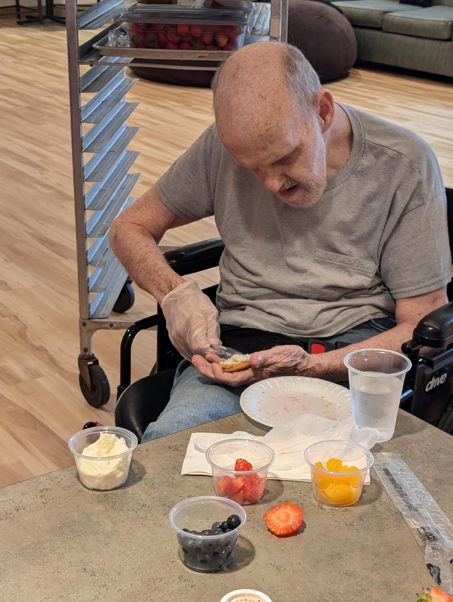 An elderly man with a bald head, wearing a gray t-shirt, seated in a wheelchair, looks down as he holds a piece of food. There is a table with various bowls of fruit and desserts, including strawberries, blueberries, and orange slices, in front of hi