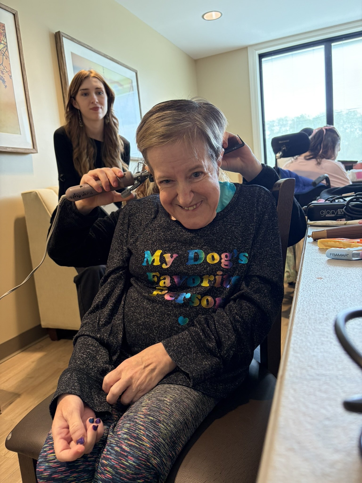 A woman with short gray hair sitting in a wheelchair smiling, while a hairdresser cuts her hair behind her. The woman is wearing a black sweater with rainbow text that says 'My Dog's Favorite' and rainbow-colored striped pants. There is another perso