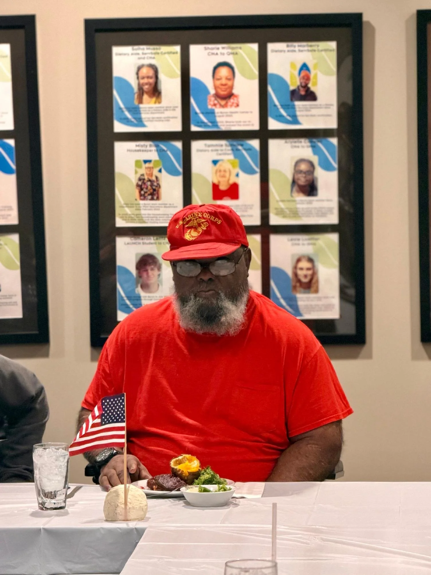 Man wearing a red shirt, red cap with yellow Marine Corps emblem, glasses, and beard sitting at a table with a white tablecloth, American flag, and food, in front of a wall with framed photo collages.