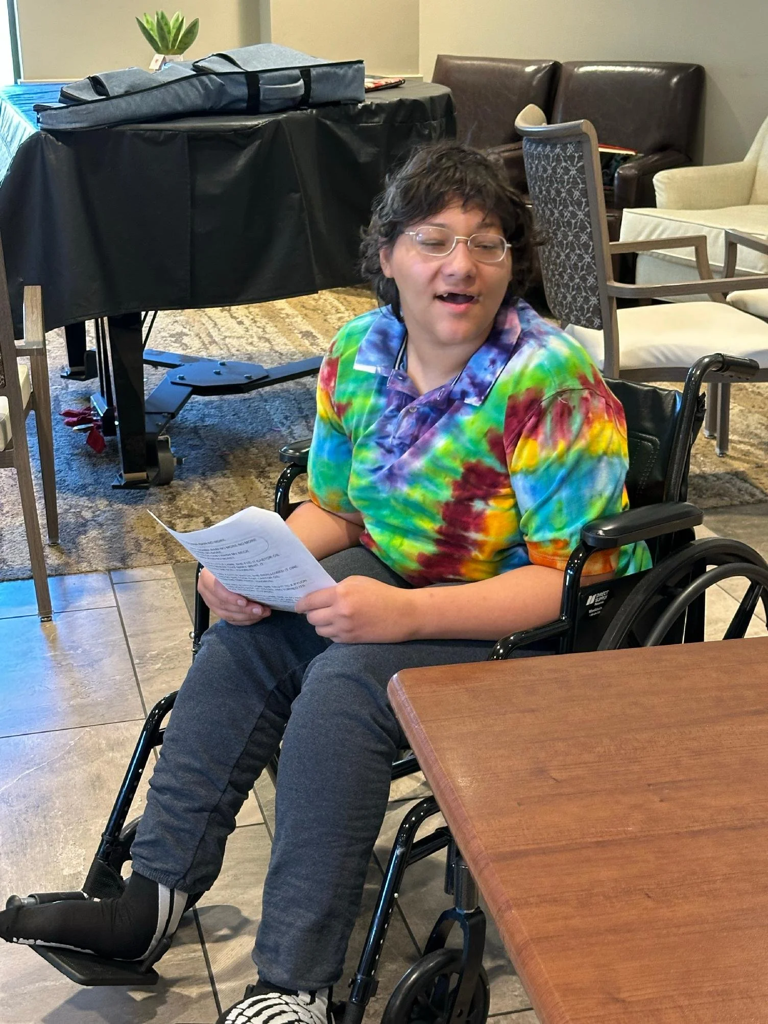 A person with short dark hair, glasses, and a tie-dye shirt sitting in a wheelchair, holding a piece of paper, in a room with chairs and tables, and a black covered table with a folded gray bag and a potted plant in the background.