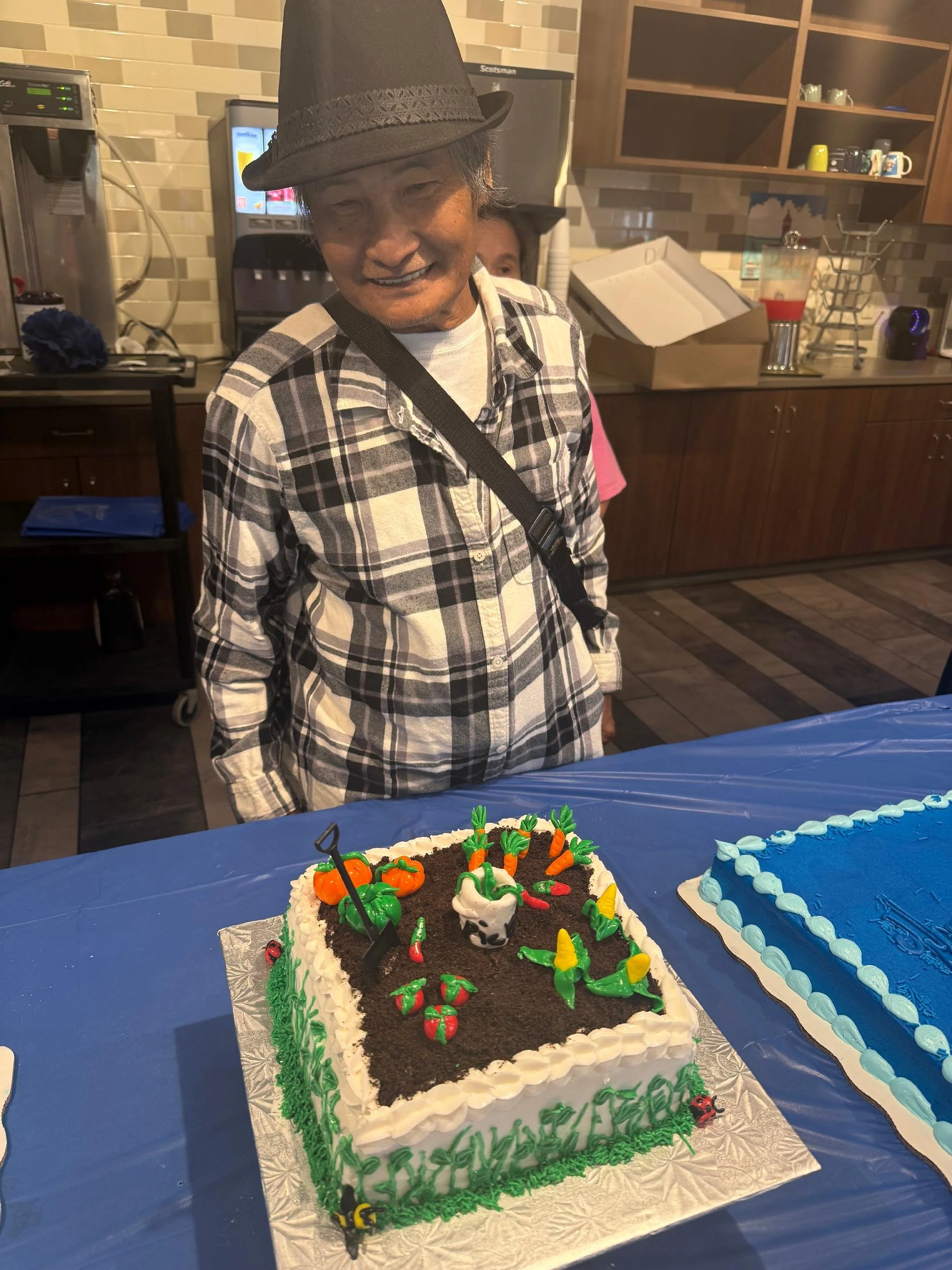 A man wearing a plaid shirt and a hat is smiling at a cake decorated with gardening theme toppers, on a table with a blue tablecloth, likely at birthday or celebration event.