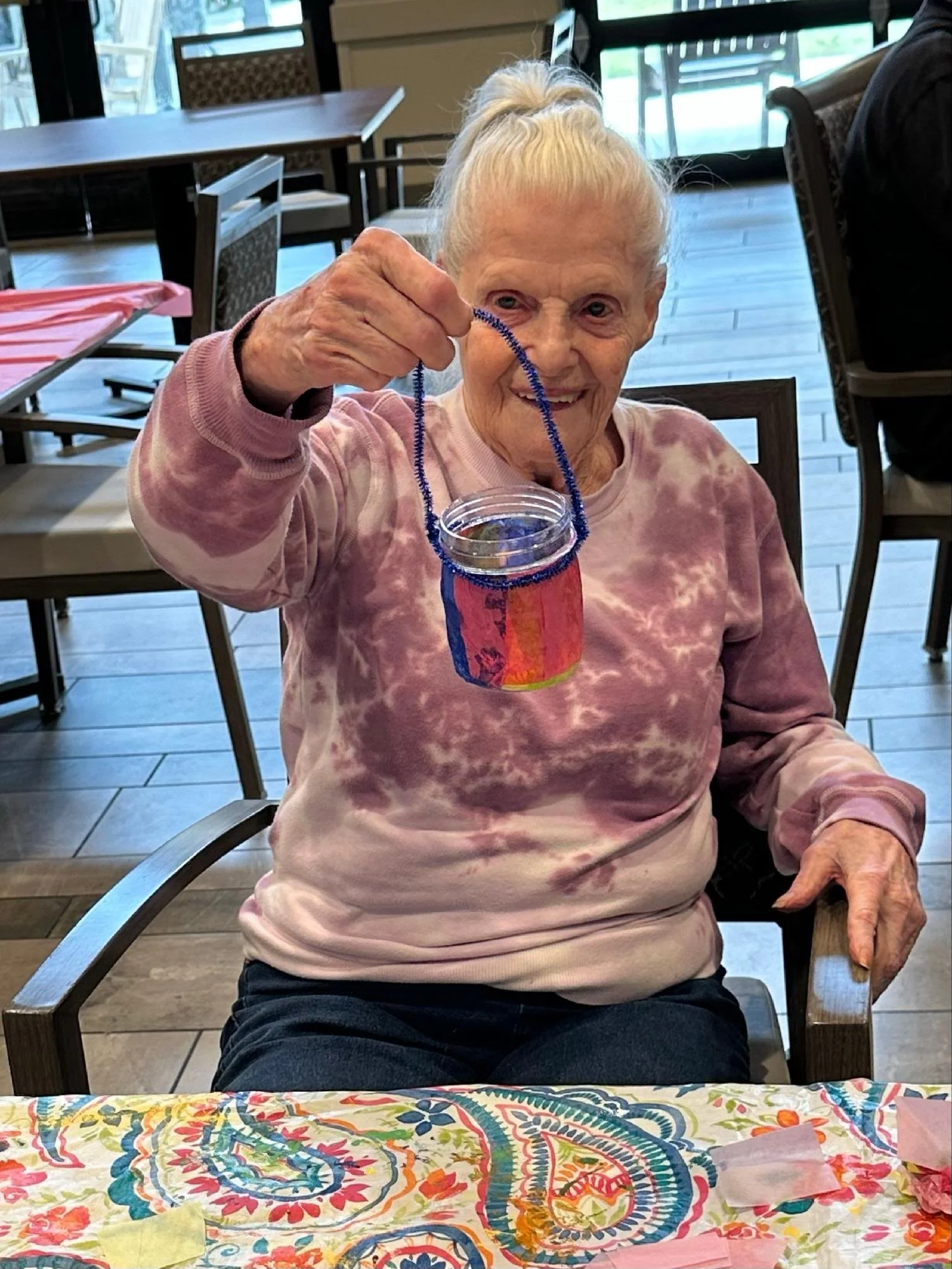An elderly woman with white hair in a ponytail smiling while holding a decorated glass jar with a blue pipe cleaner handle, sitting in a wooden chair at a dining table with a colorful tablecloth and paper decorations. The background shows glass doors