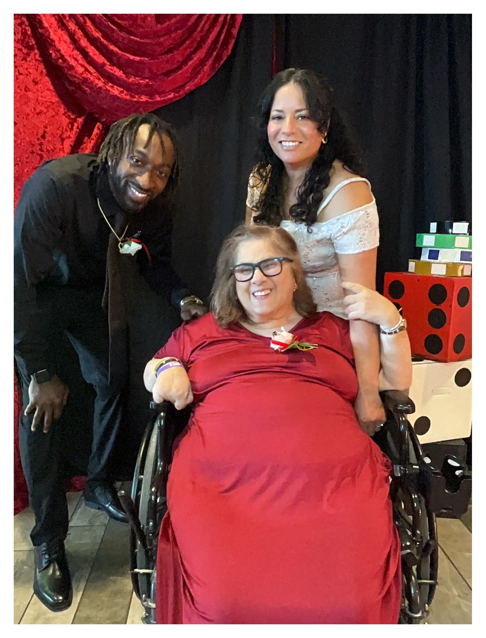 Four women, one in a wheelchair, are posing together in front of a backdrop with red and black curtains. The woman in the wheelchair is wearing a red dress and has a red rose pinned to her chest. The woman standing to her left is wearing black and ha