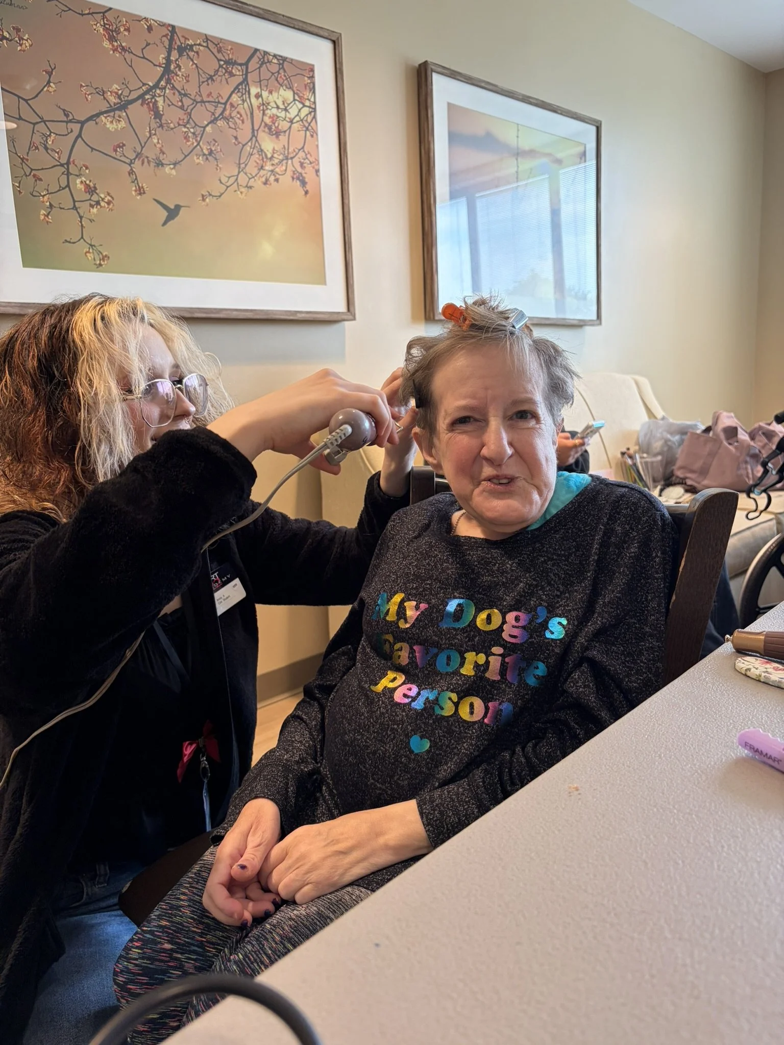 An elderly woman with short gray hair and a colorful shirt that reads 'My Dog's Favorite Person' is getting her hair done by a young woman with glasses and curly blonde hair. The young woman is using a hairdryer. They are sitting at a table in a room