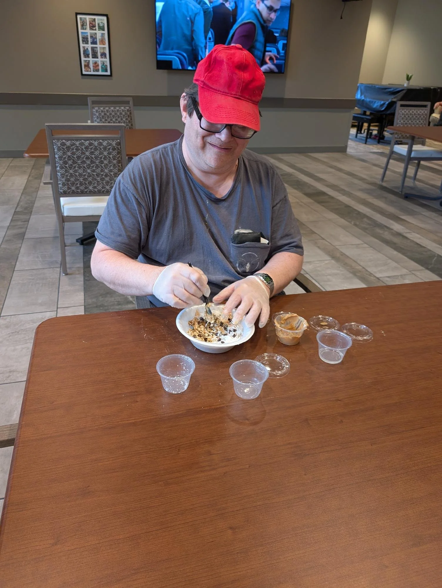A man with glasses, wearing a red cap and gray t-shirt, is sitting at a wooden table and scooping ice cream or dessert from a bowl. There are small clear plastic cups and containers with sauce or toppings on the table. The background shows a televisi