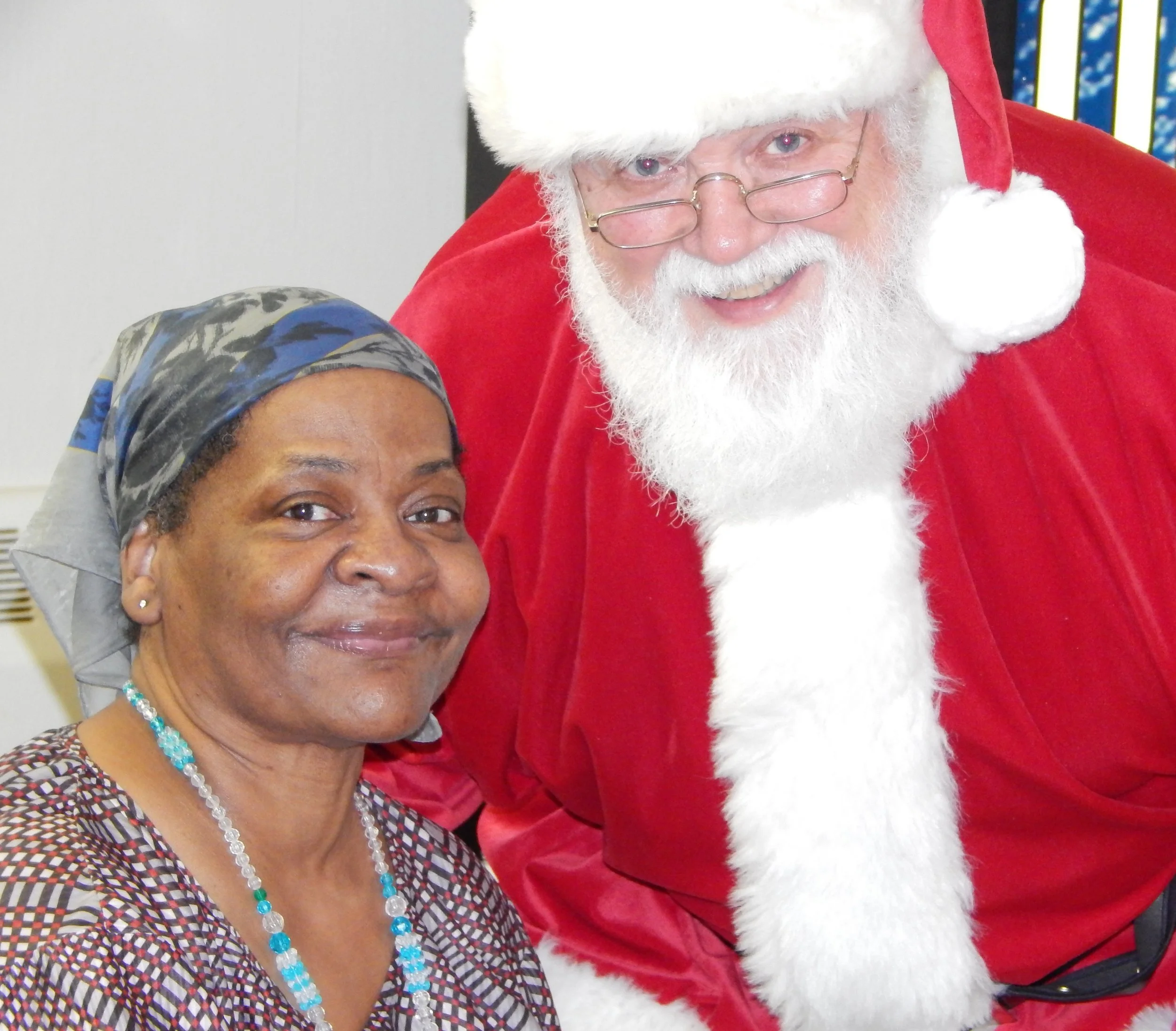 A woman with dark skin and a patterned dress is taking a selfie with Santa Claus, who is dressed in his traditional red suit and white beard, smiling at the camera.