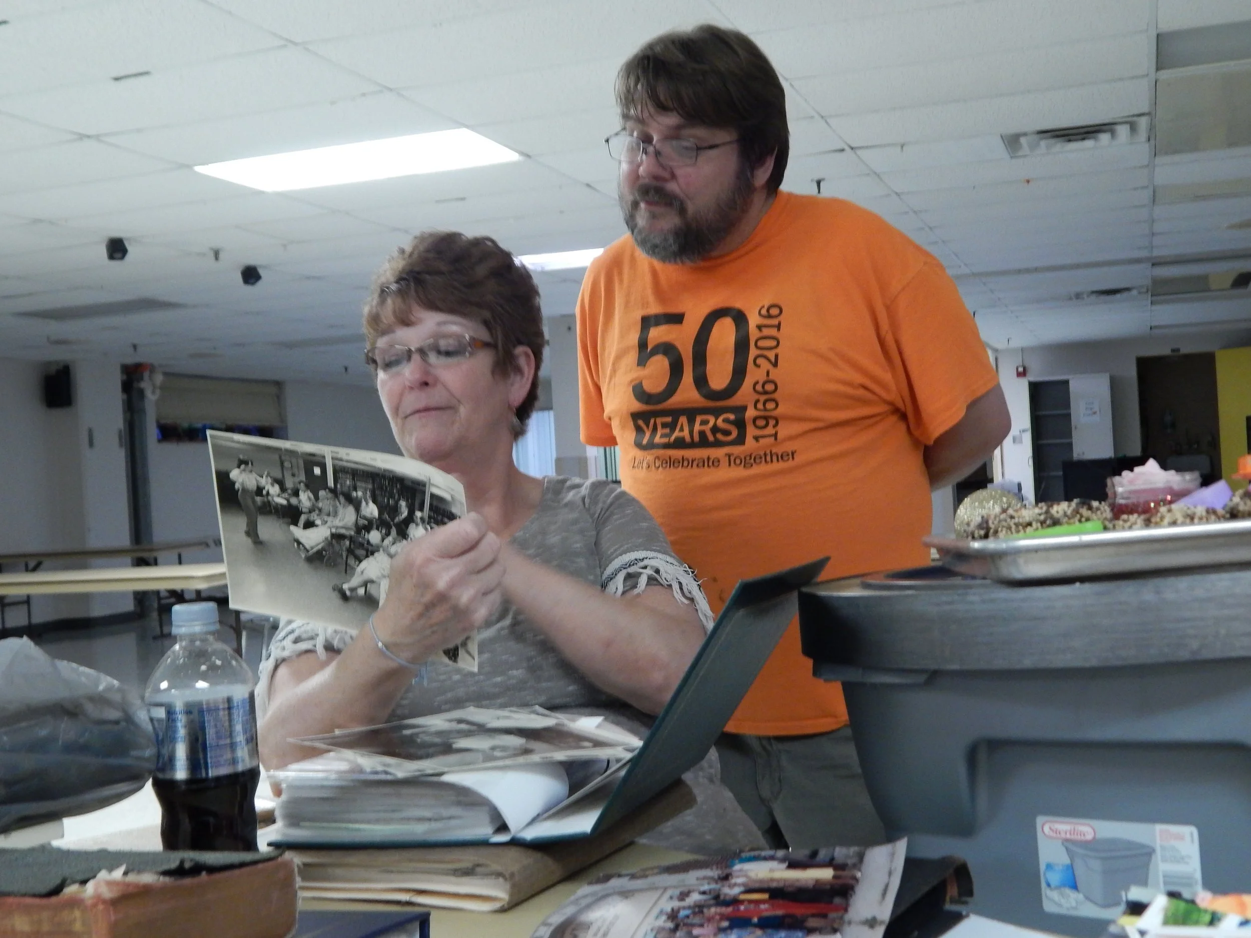 A woman is sitting at a table looking at a black-and-white photograph. A man standing behind her wears an orange shirt celebrating 50 years at an event.