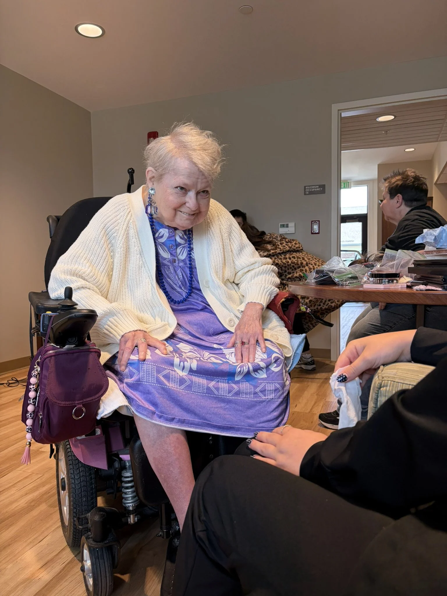 An elderly woman in a wheelchair getting her blood drawn by a healthcare professional in a medical facility. She is smiling and looking at the person administering the test, with other people in the background.