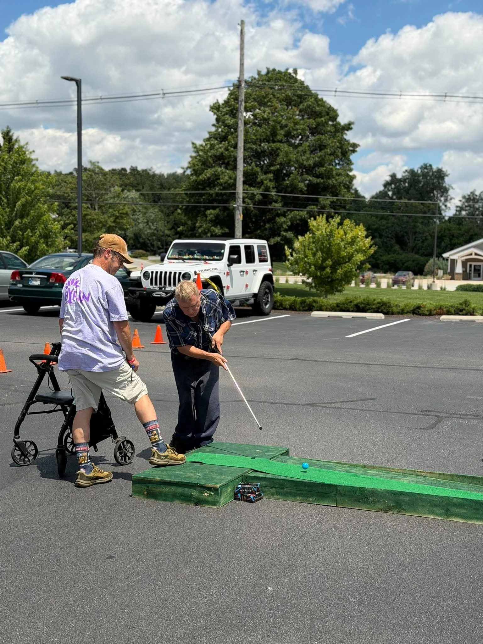 Two men are playing mini golf outdoors in a parking lot, with one man bending down and the other standing next to a golf ball on a green mat, surrounded by orange cones and a green mini golf hole. There are cars parked beside them and trees with a pa