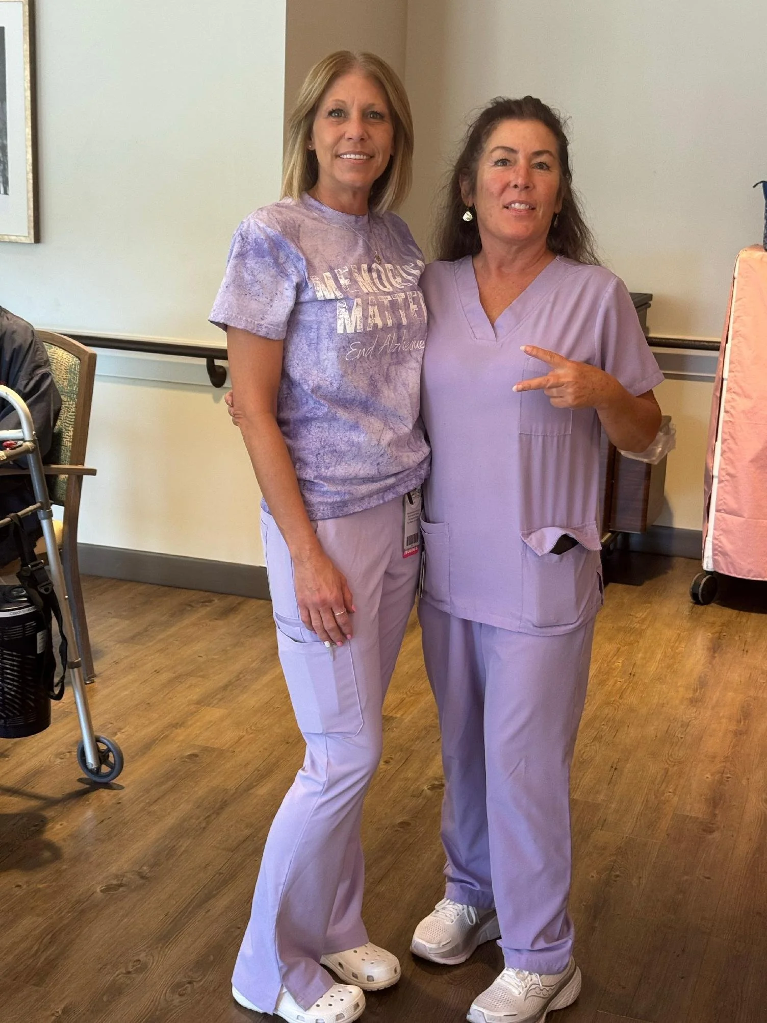 Two women wearing medical scrubs and sneakers standing inside a room, smiling at the camera. One is pointing at the other.