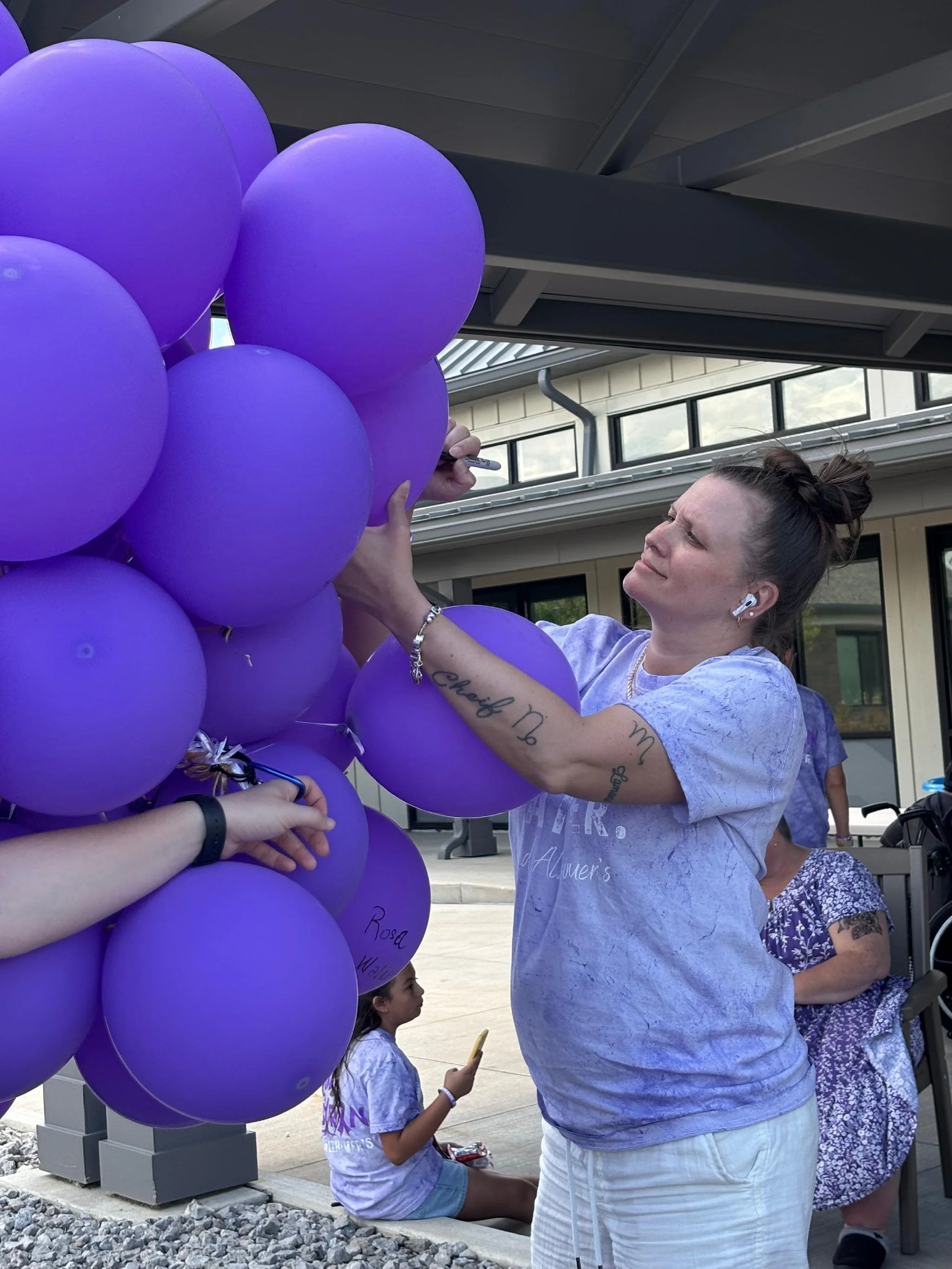 A woman with tattoos on her arm and wearing earbuds is signing purple balloons with words. Several children and adults are nearby, and they are outdoors near a building with windows and a roof overhang.